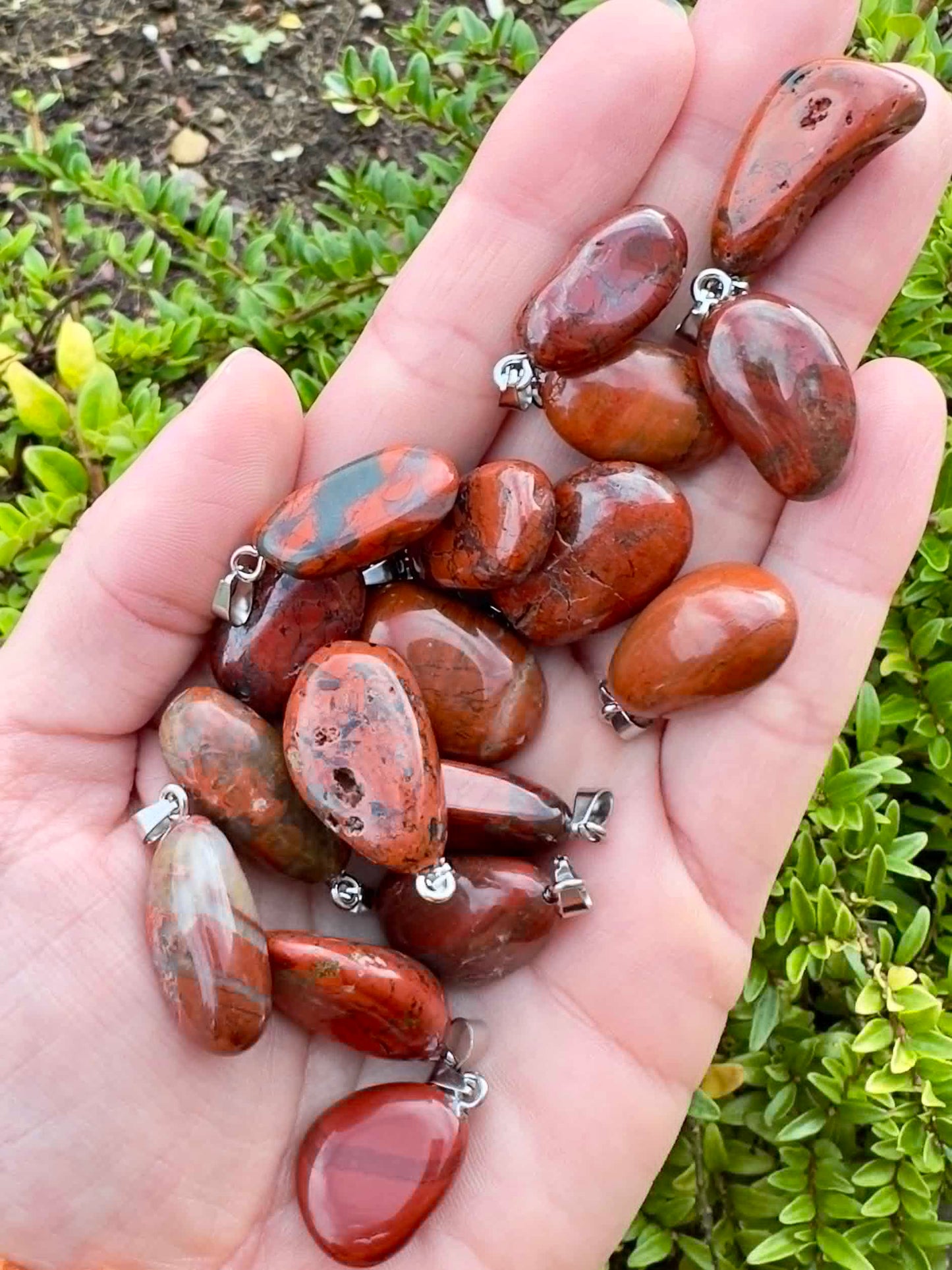 Hånd fuld af Rød Jaspis vedhæng, foran grønne blade i baggrunden.