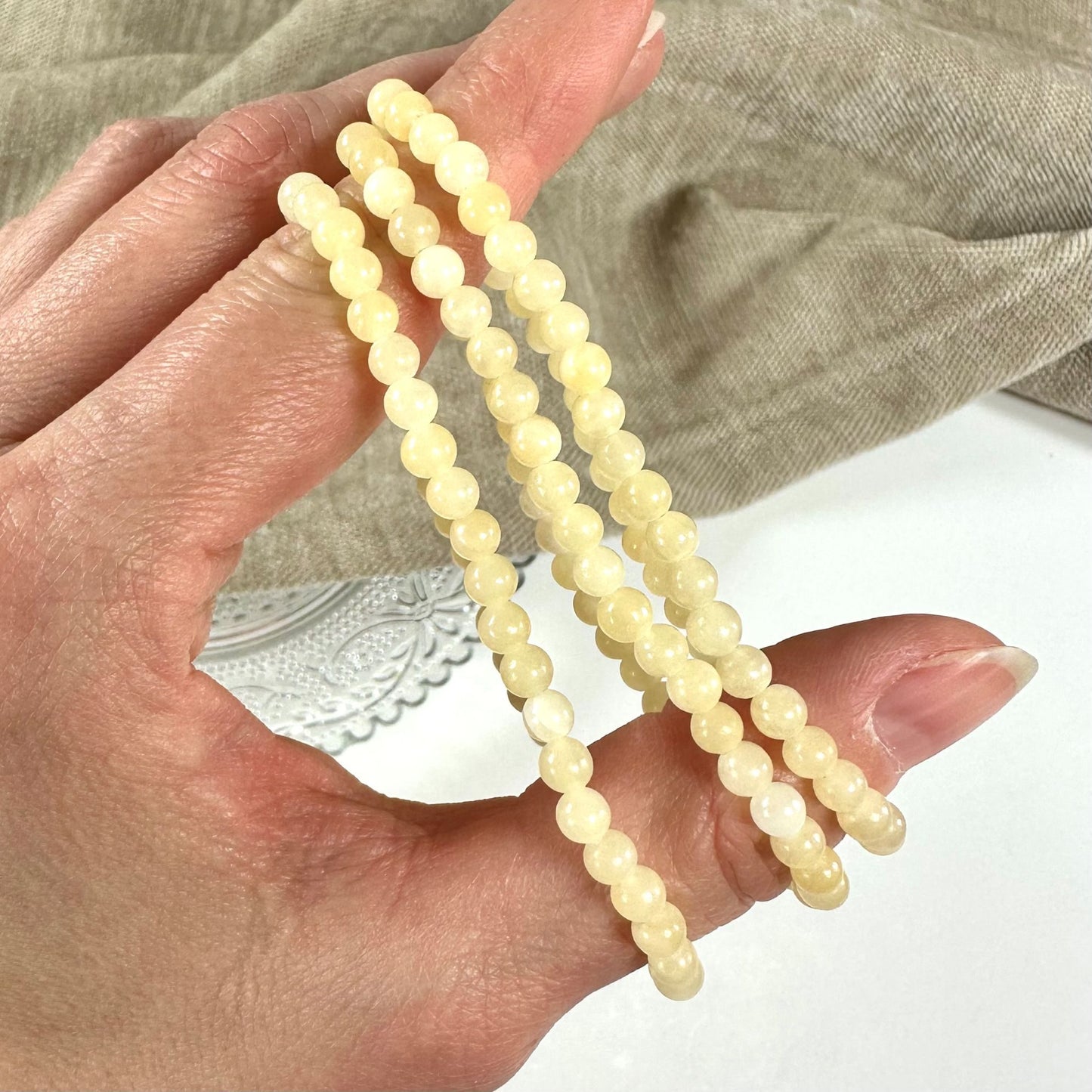 Hånd der holder tre gul calcit armbånd med perler, baggrund af beige stof.
