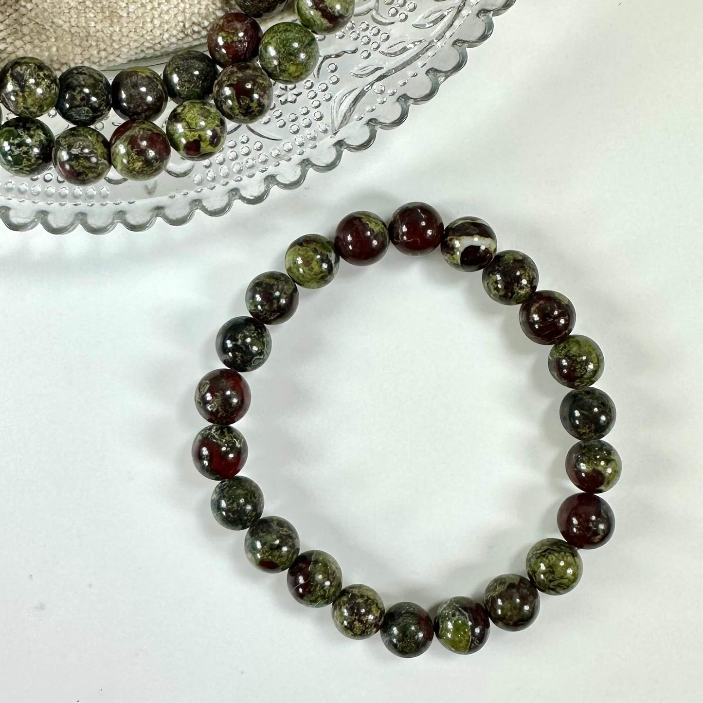 Drageblod armbånd med grønne og mørkerøde perler, ligger på en hvid baggrund ved siden af flere perler.