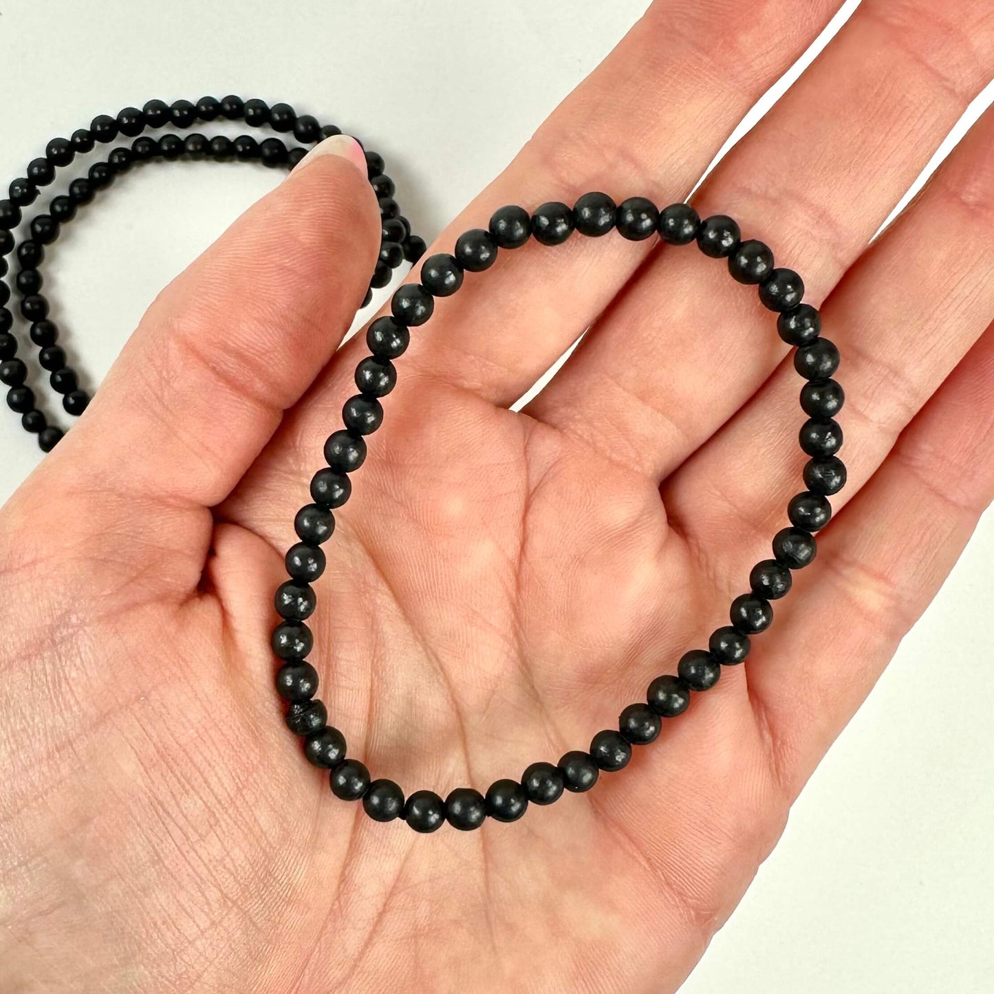 En hånd holder et shungit armbånd lavet af sorte perler, med et lignende armbånd i baggrunden.