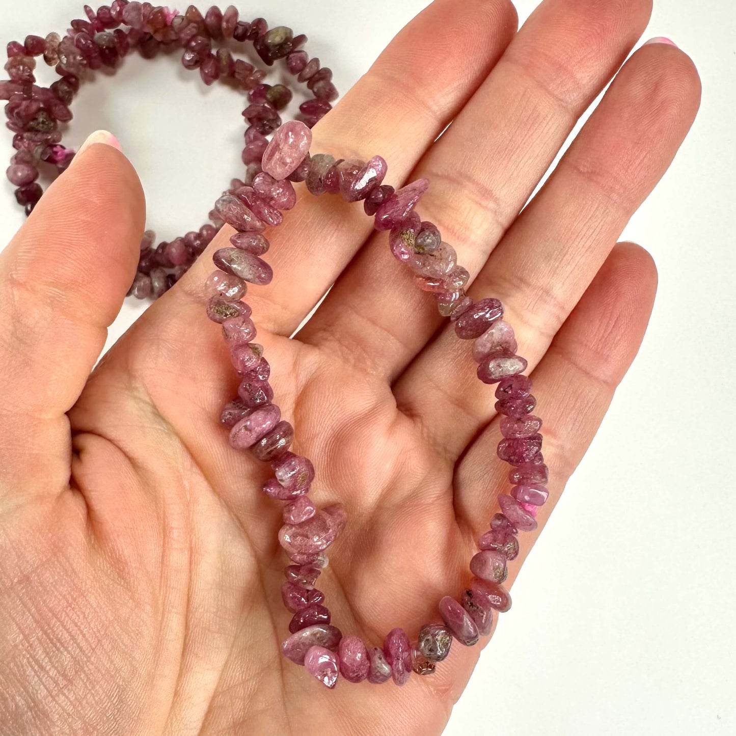 En hånd holder et pink turmalin armbånd, bestående af uregelmæssige perler. Et andet armbånd ses i baggrunden.