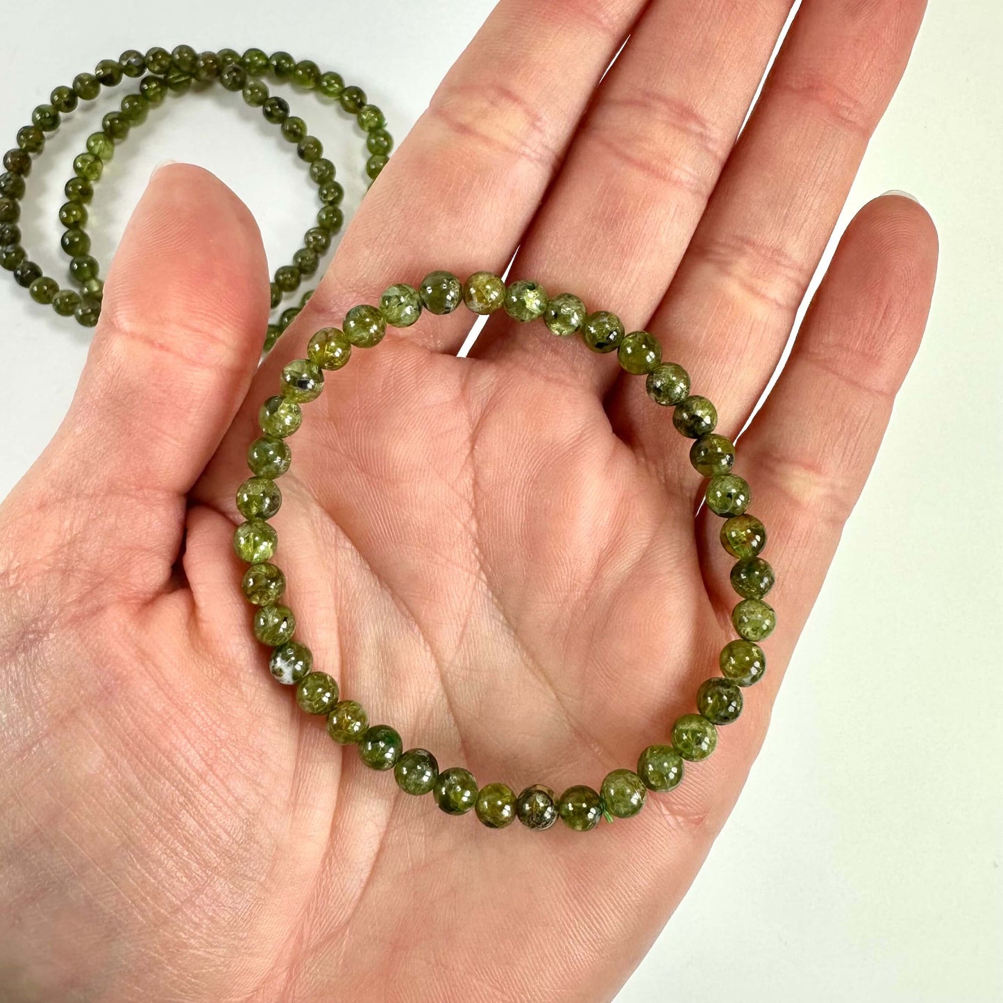 En hånd holder et grønt Peridot armbånd, med to lignende armbånd i baggrunden.