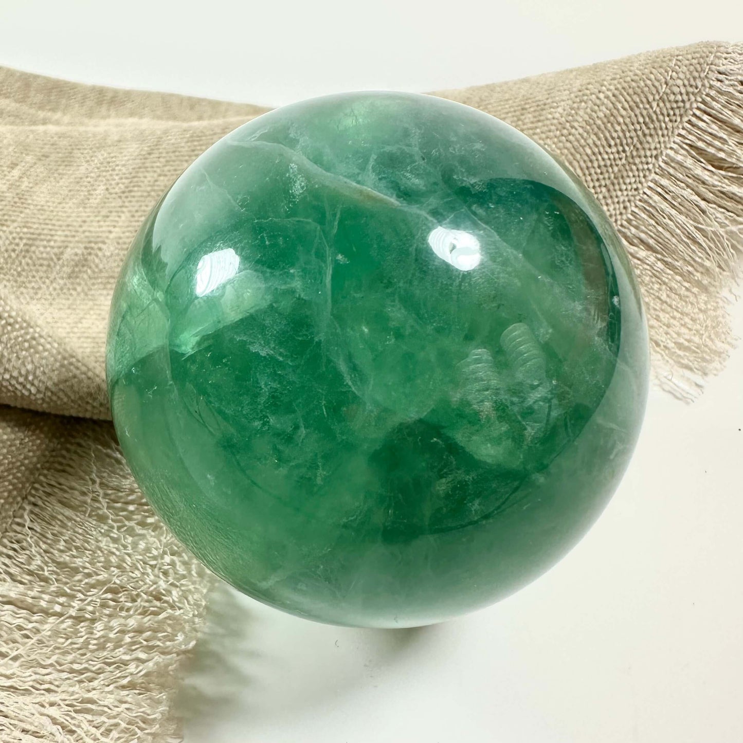 Grøn fluorit kugle på en beige klud med frynser, fotograferet mod en neutral baggrund.