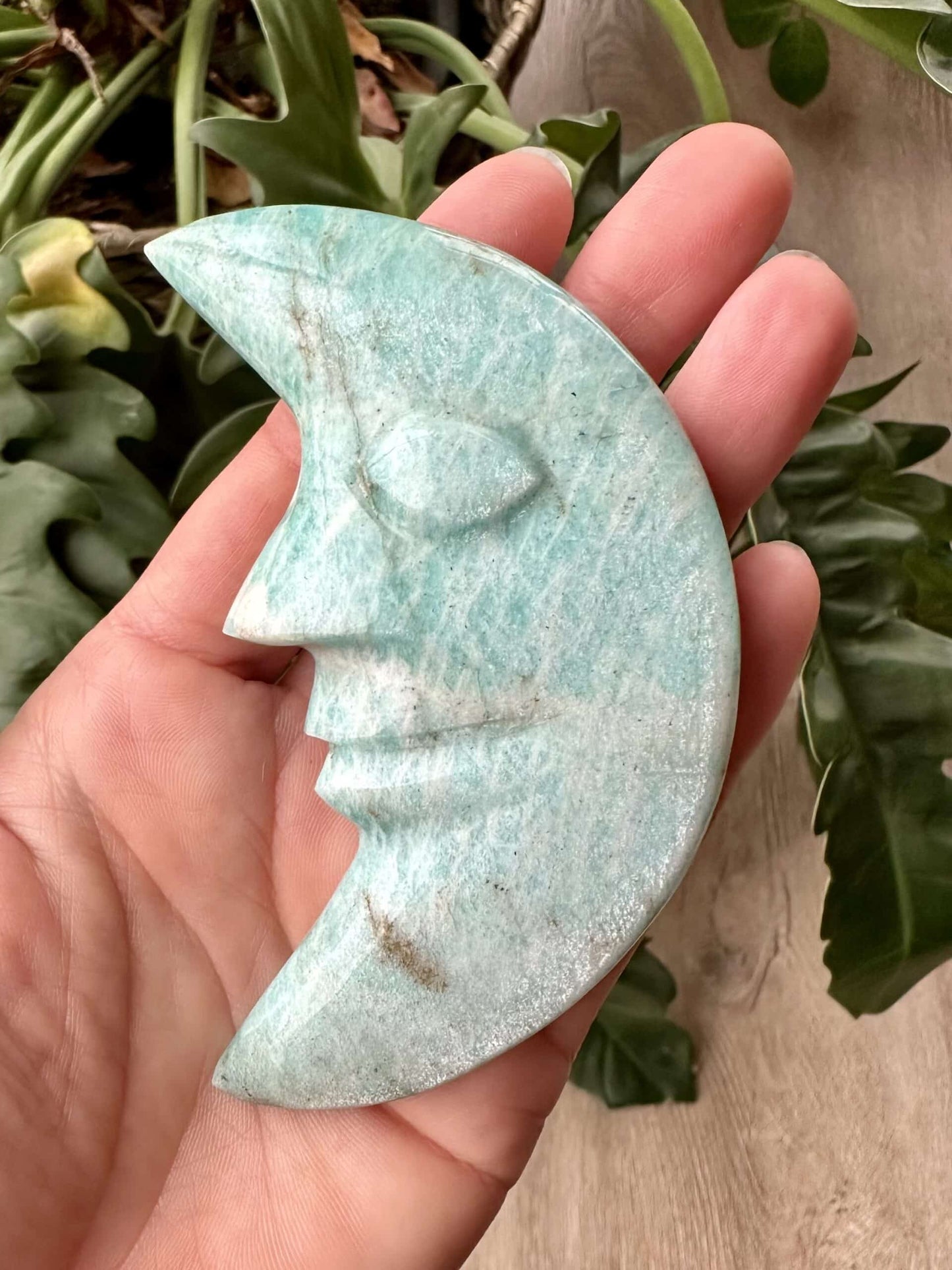 Hånd holder en halvmåneformet skulptur lavet af amazonit, med detaljeret ansigt, omgivet af grønne blade.