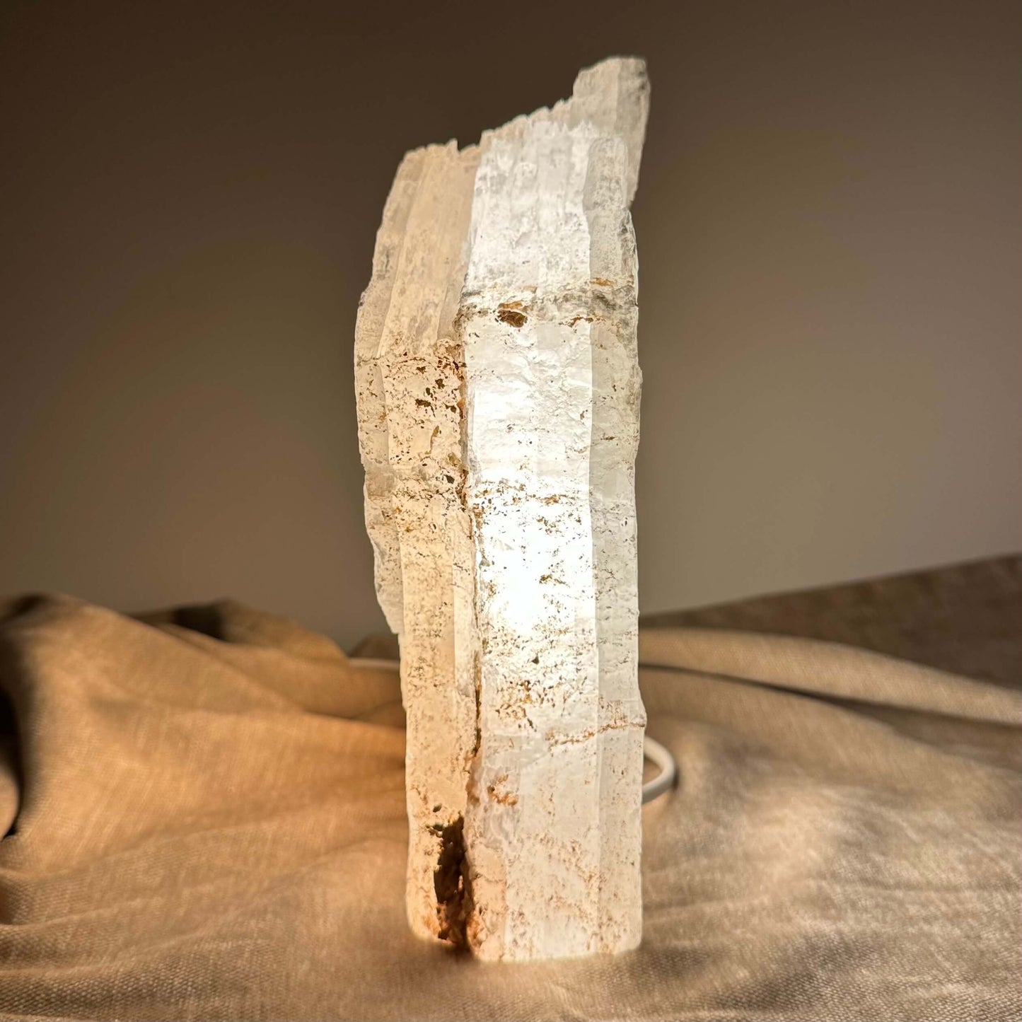 En tændt selenitlampe står på en beige tekstilbaggrund, der skaber en varm og rolig stemning.