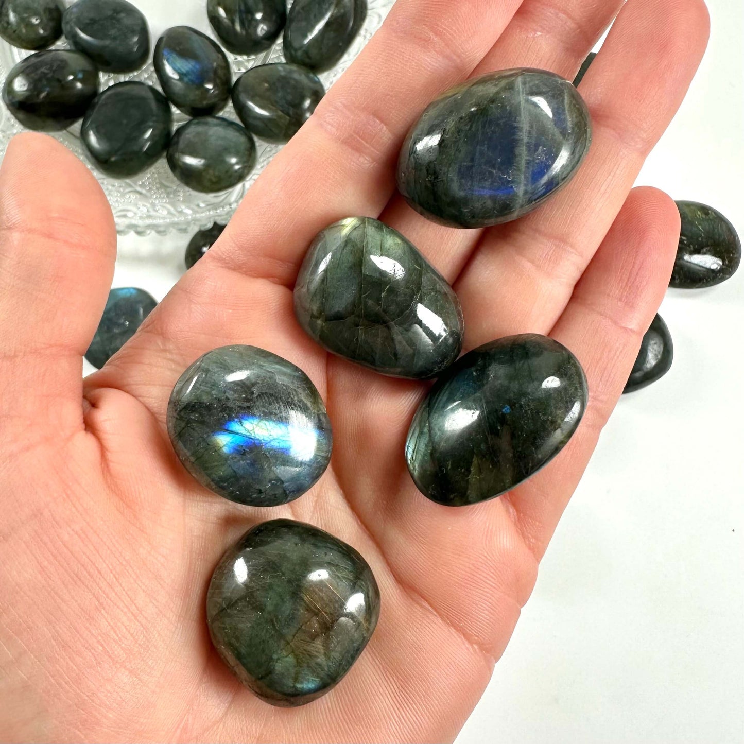 En hånd holder flere glatte Labradorit lommesten, med en skål af lignende sten i baggrunden.