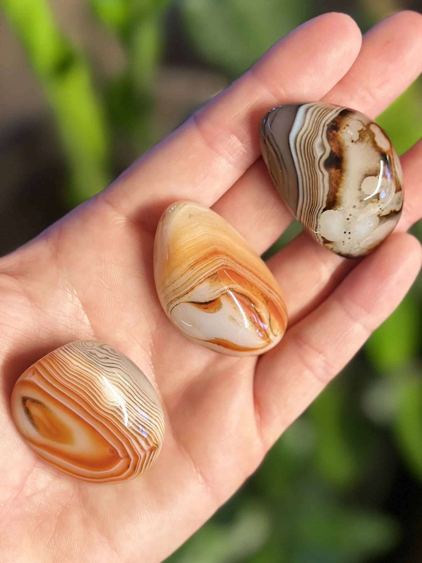 En hånd holder tre sardonyx lommesten med karakteristiske båndmønstre i nuancer af brun, orange og hvid.