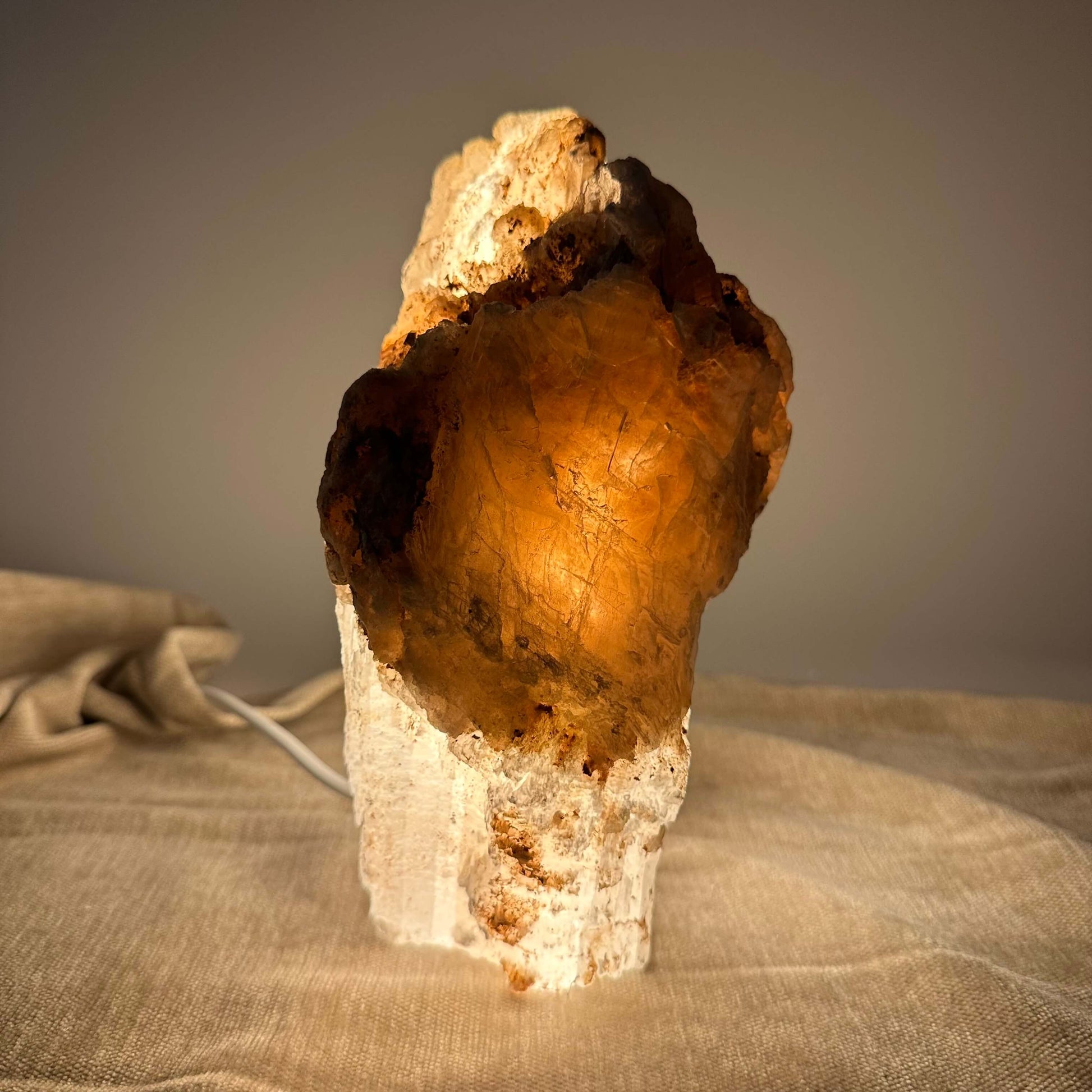 Mexicansk Selenit lampe nr 6 lyser blødt op med en varm, ravfarvet glød på et beige stofunderlag.