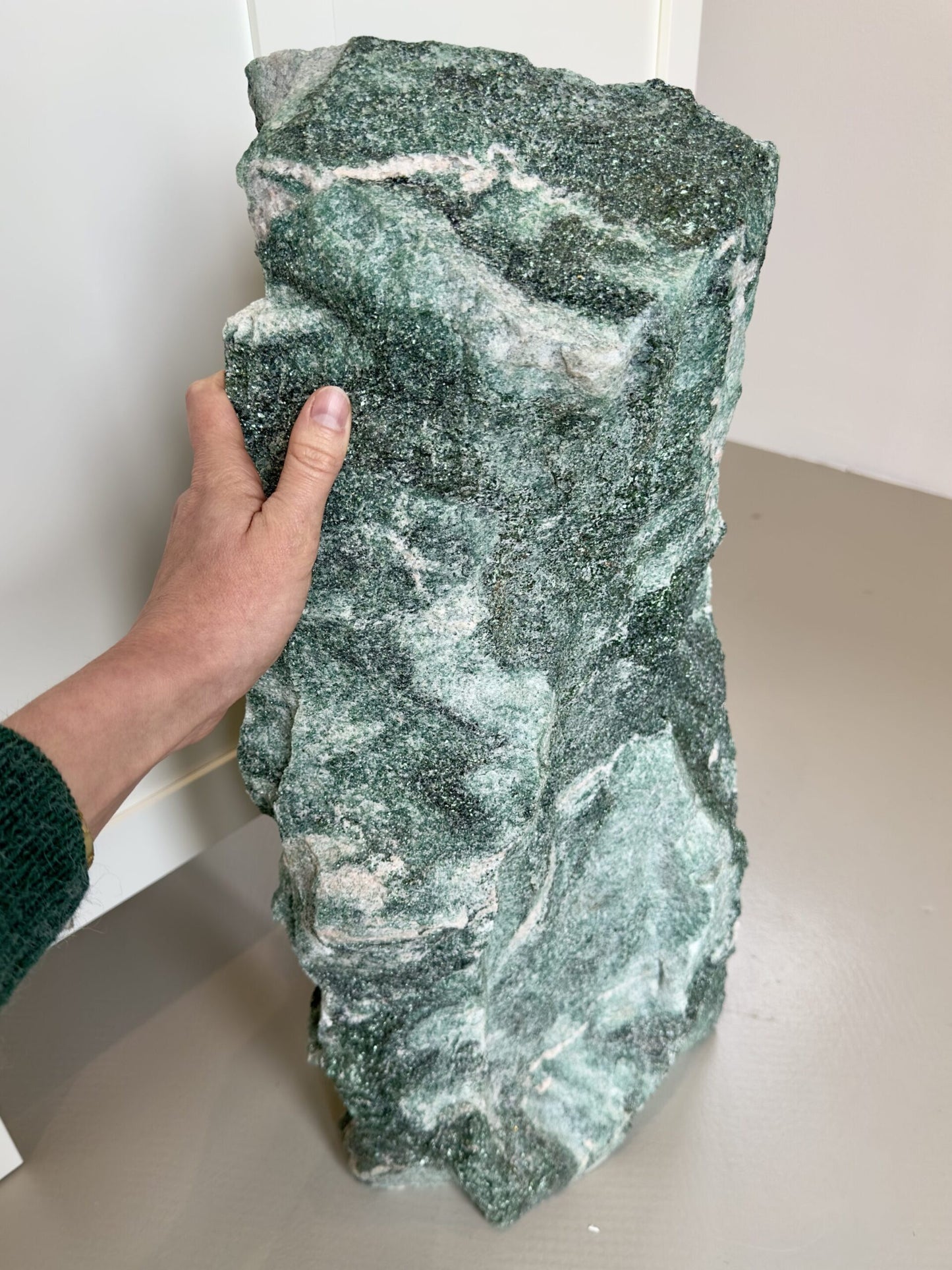 Hånd holder stor, flad, grøn sten med hvidlige årer på en beige baggrund.