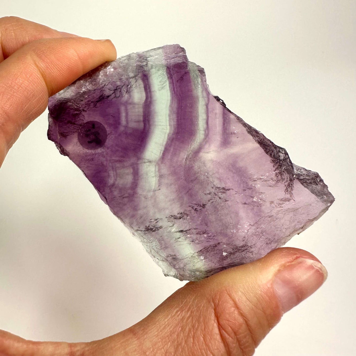 Hånd der holder en regnbue fluorit skive med lilla og hvide striber mod en lys baggrund.