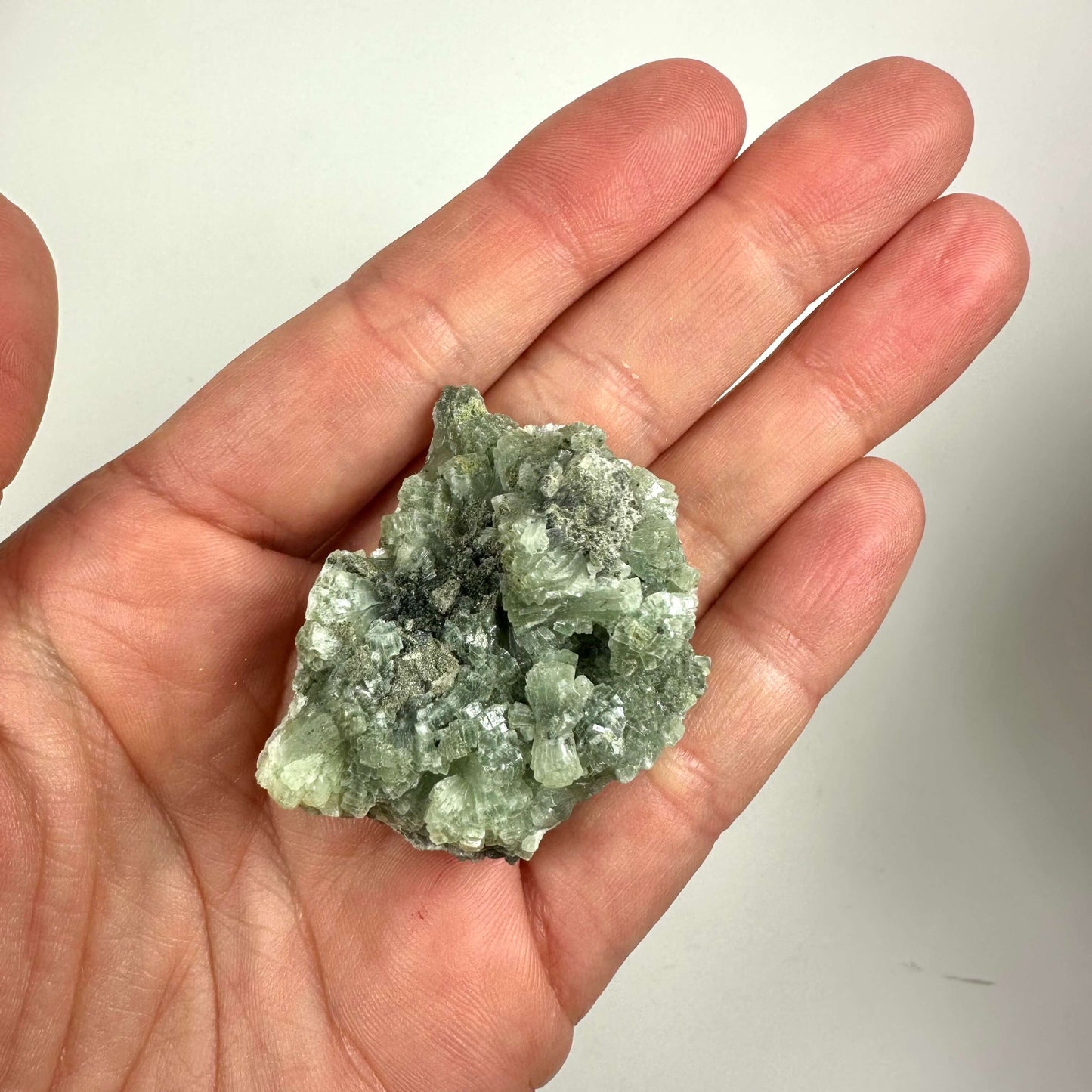 Hånd holder en grøn Prehnit klynge krystal på en lys baggrund.