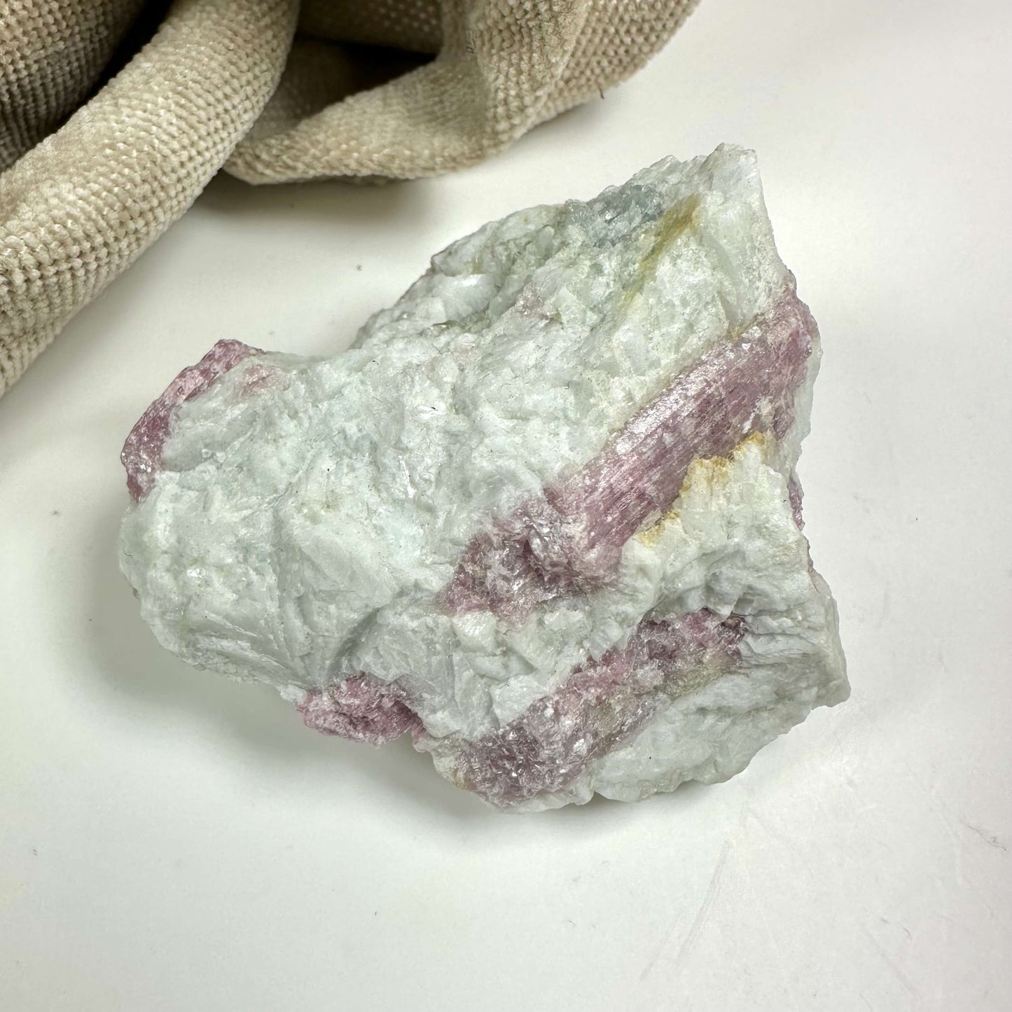 Rå rubellit kvarts med lyserøde og hvide nuancer placeret på en lys overflade ved siden af en beige stofpose.