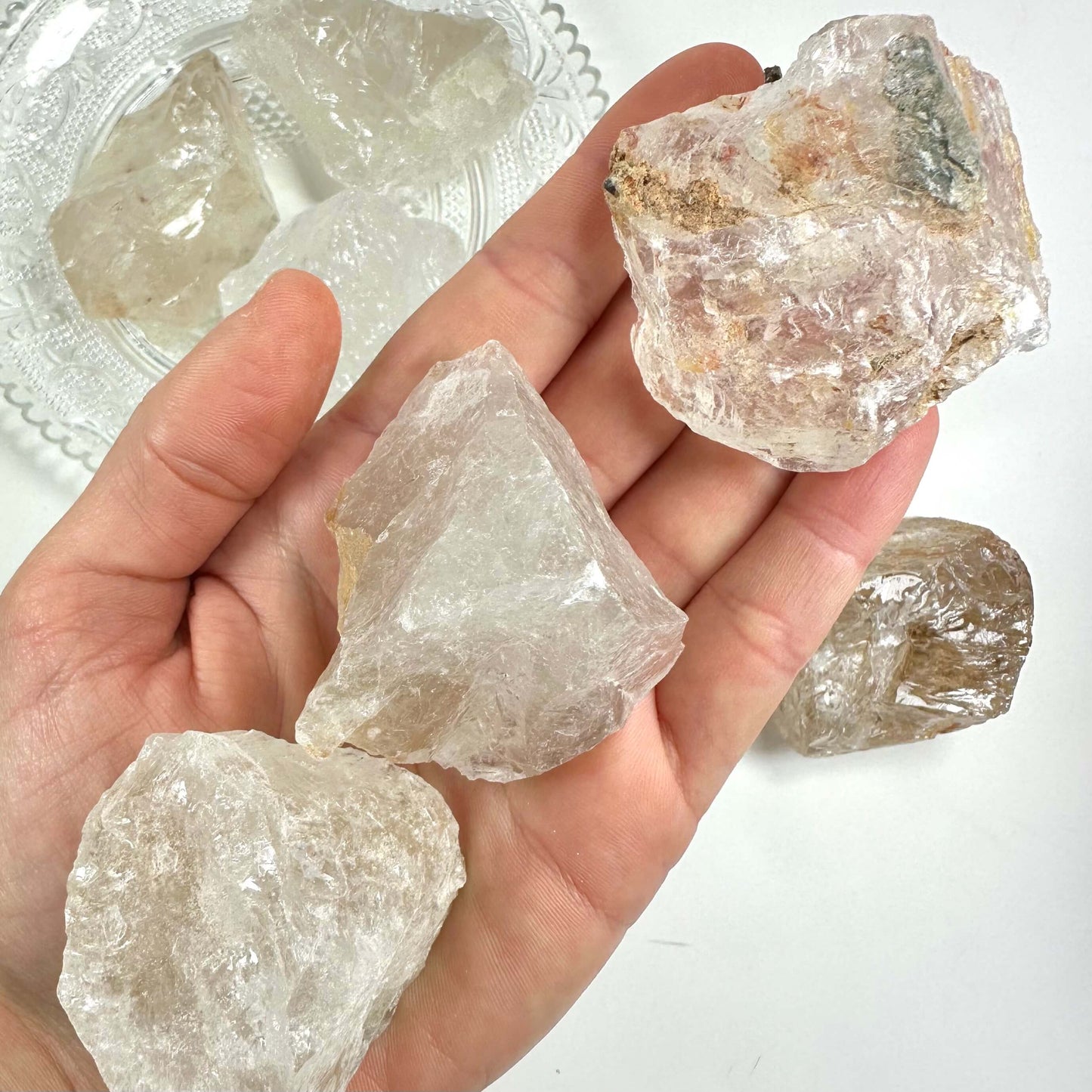 Hånd holder flere store, rå bjergkrystaller mod en hvid baggrund. Flere krystaller er på en tallerken i baggrunden.