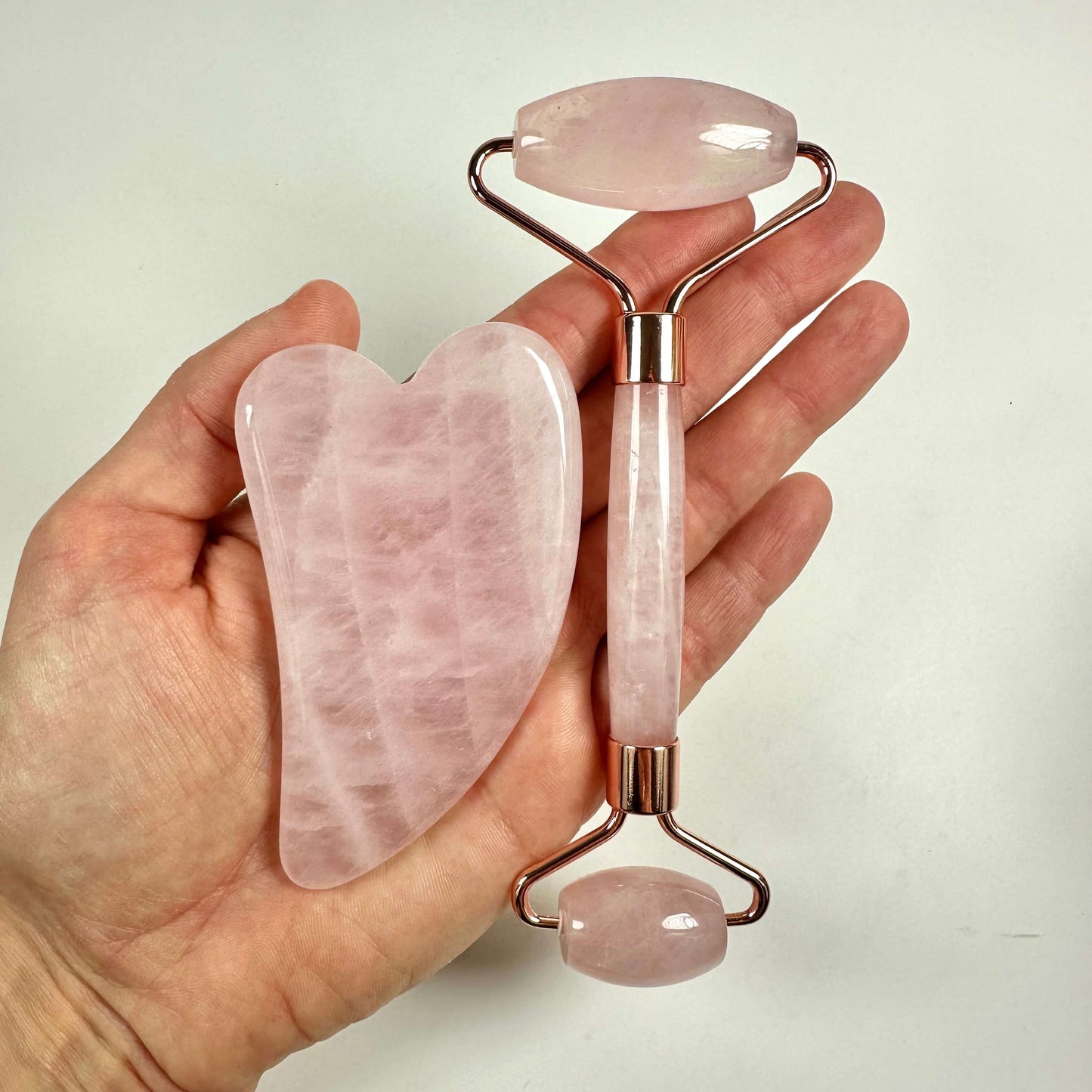 Hånd holder et Gua sha sæt med en lyserød rulle og hjerteformet sten på hvid baggrund.
