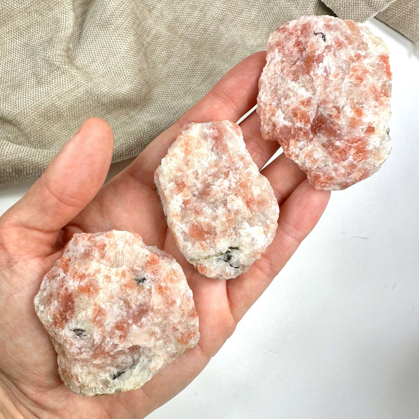 En hånd holder tre stykker solsten rå, med rosa og hvide nuancer på en lys baggrund.