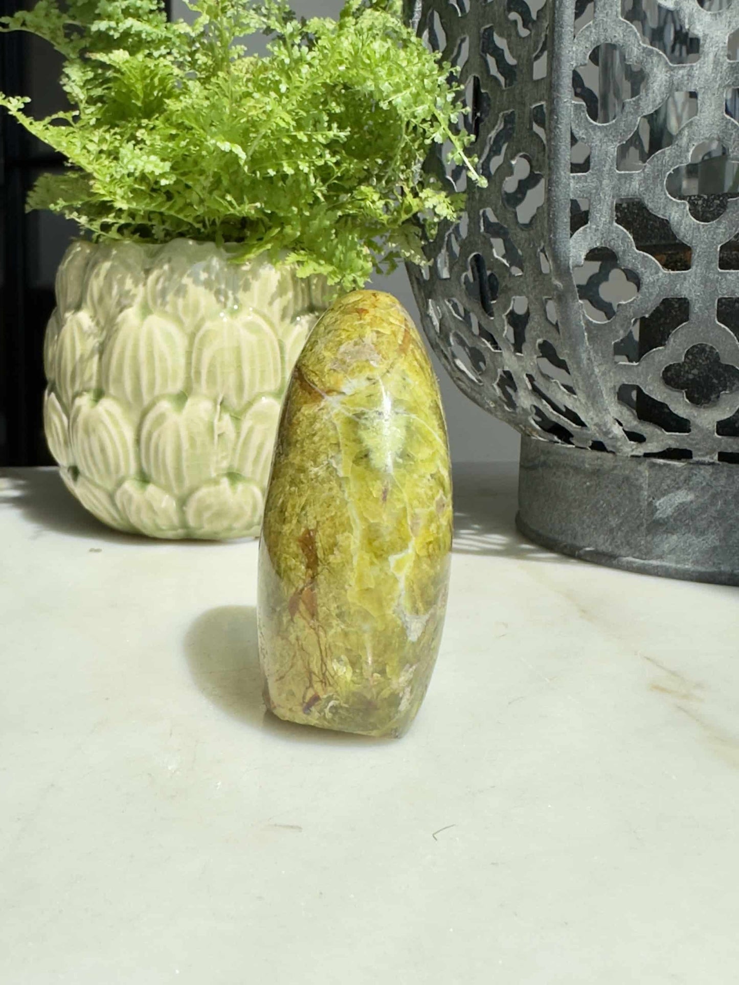 Grøn opal sten i fri form på bord, med grøn plante og metaldekoration i baggrunden.