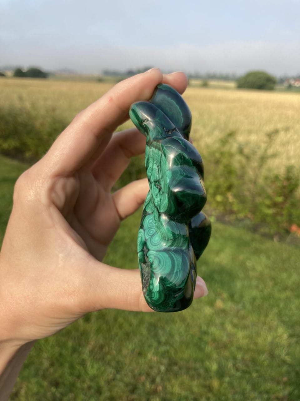 Hånd holder en grøn malakitskulptur i sollys med mark i baggrunden.