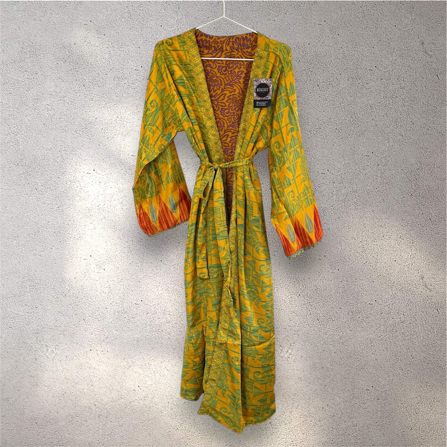 Gul og grøn mønstret kimono med røde og blå ærmer, hængende på en grå væg.