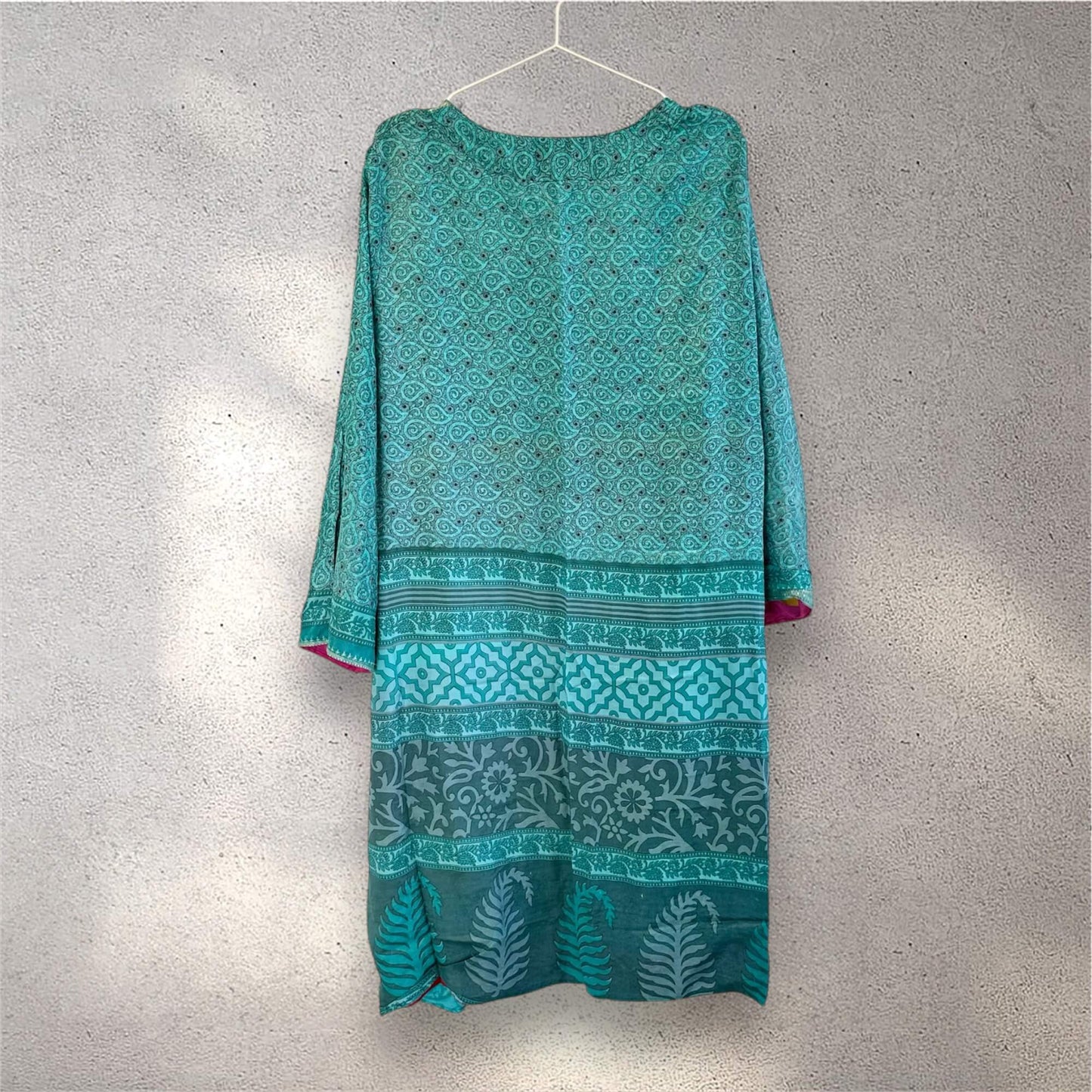 Turkis kimono med mønstre, hænger på en bøjle mod en grå væg.