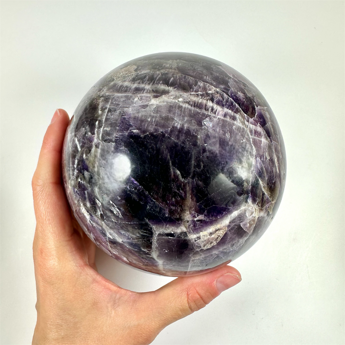 Hånd holder en stor, poleret ametyst kugle med mørke lilla nuancer på en hvid baggrund.
