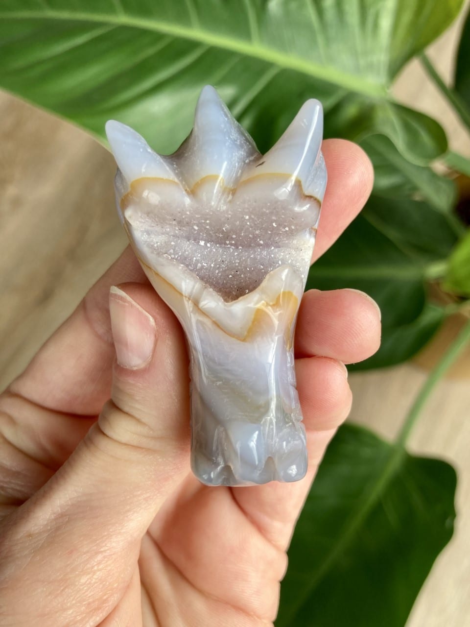 Hånd holder en dragefigur udskåret i agat natursten med grønne blade i baggrunden.
