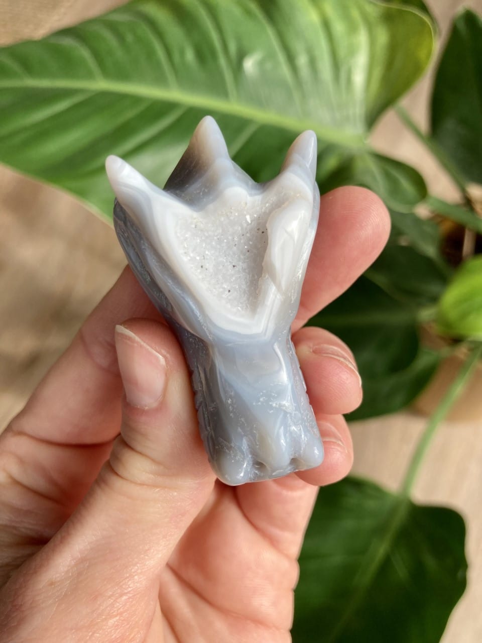 En hånd holder en skulpturel drageformet agat med naturfarver og krystalstruktur. Grønt blad i baggrunden.