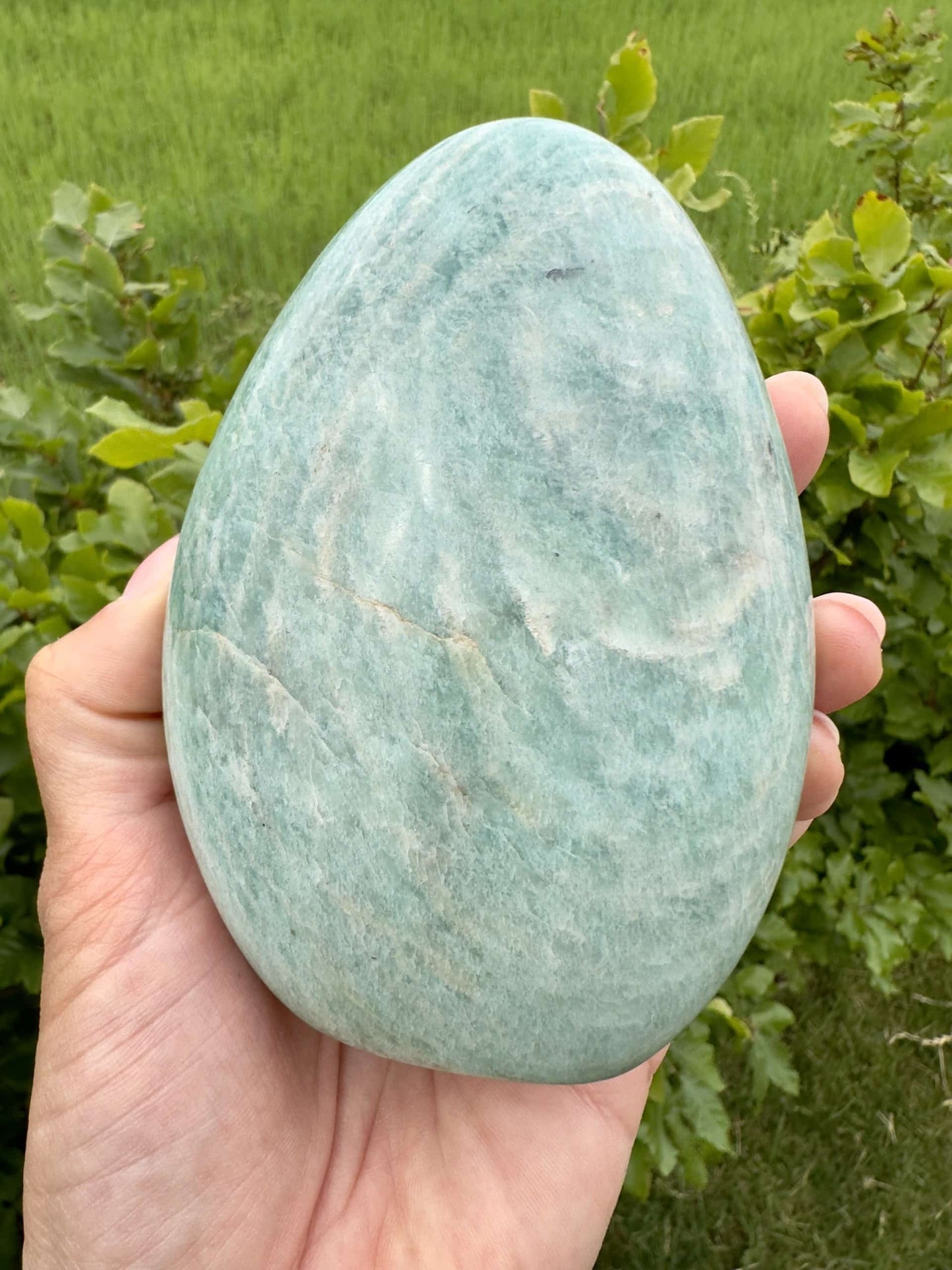 Hånd holder en stor, glat amazonit-sten i naturligt lys med grønne planter i baggrunden.