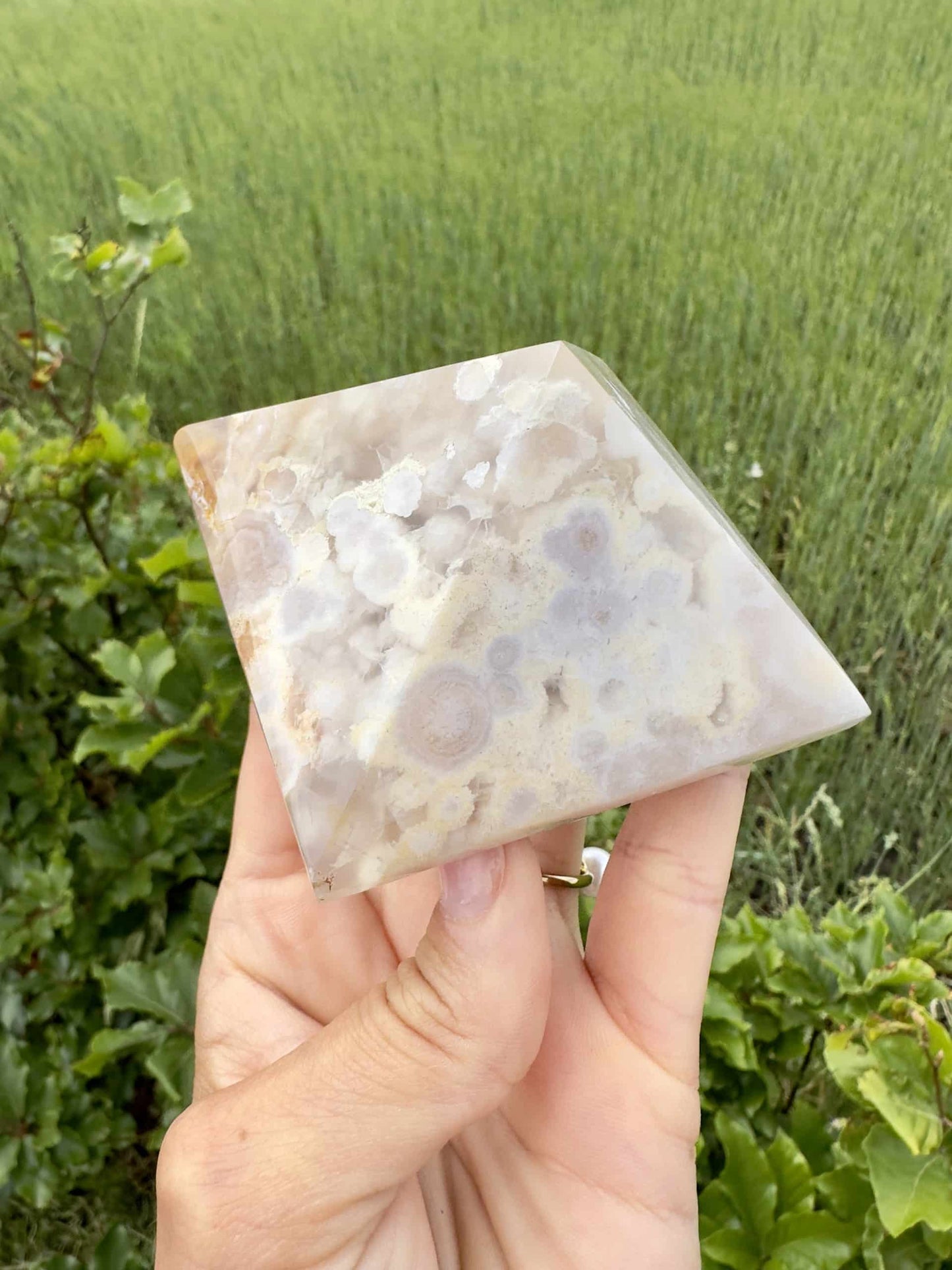 En hånd holder en Blomster agat pyramide foran en grøn græsmark.