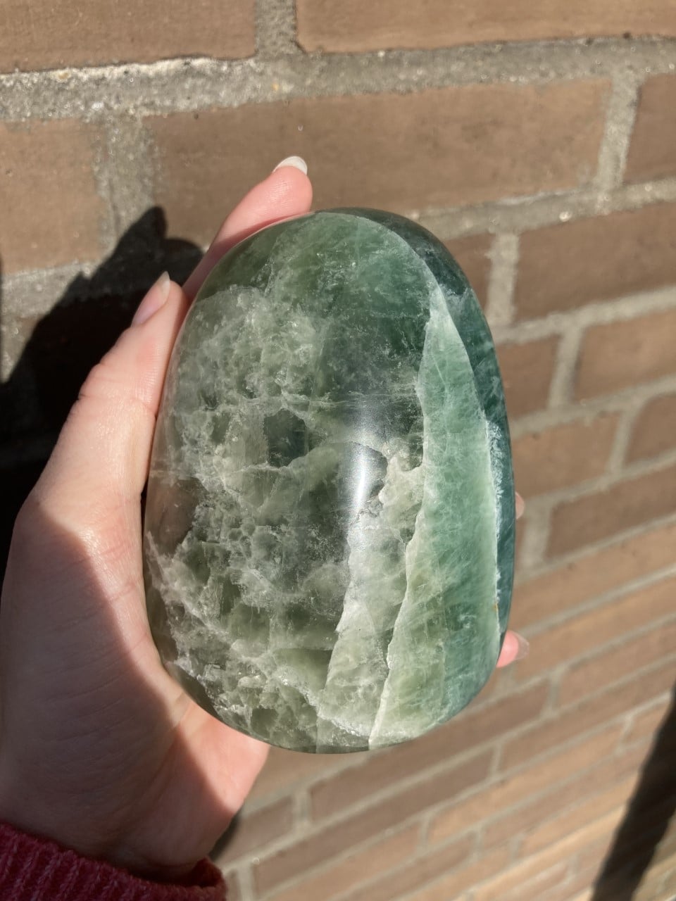 Hånd holder en glat, poleret fluorit-sten foran en murstensvæg i sollys.