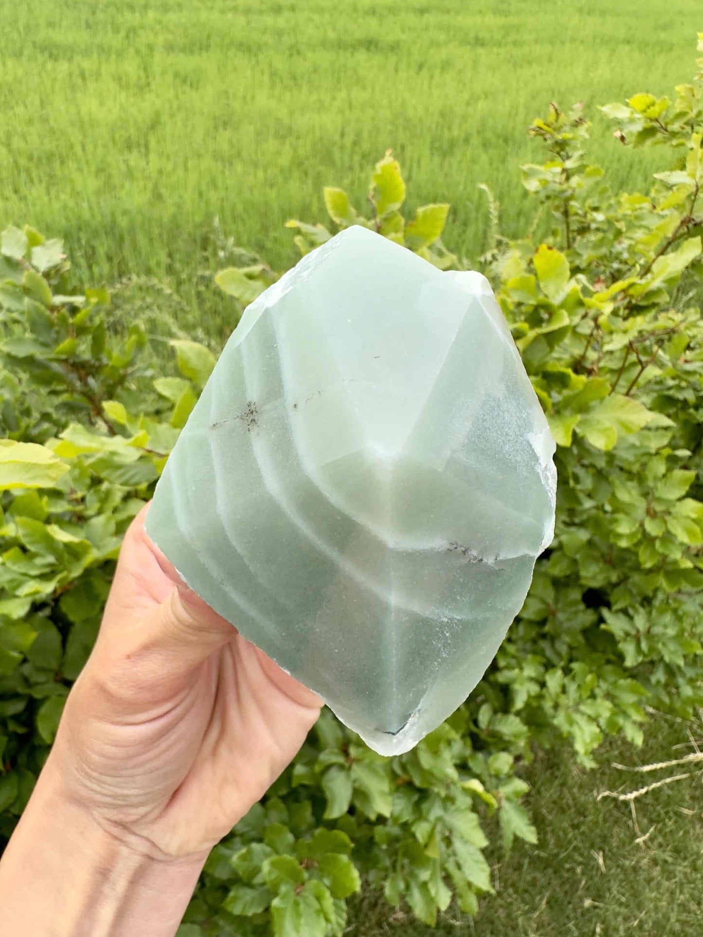 Hånd holder en stor Grøn Aventurin spids foran en grøn busk og mark i baggrunden.