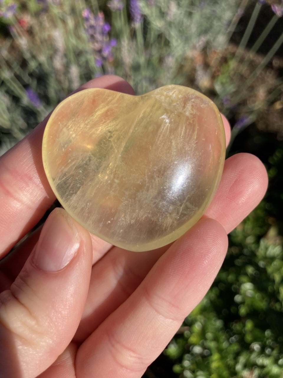 Hånd, der holder et gul fluorit hjerte, med en uskarp naturbaggrund.