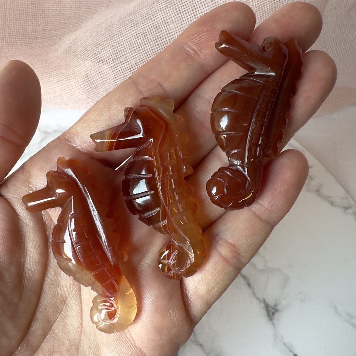 En hånd holder tre udskårne karneol søheste mod en lys baggrund.