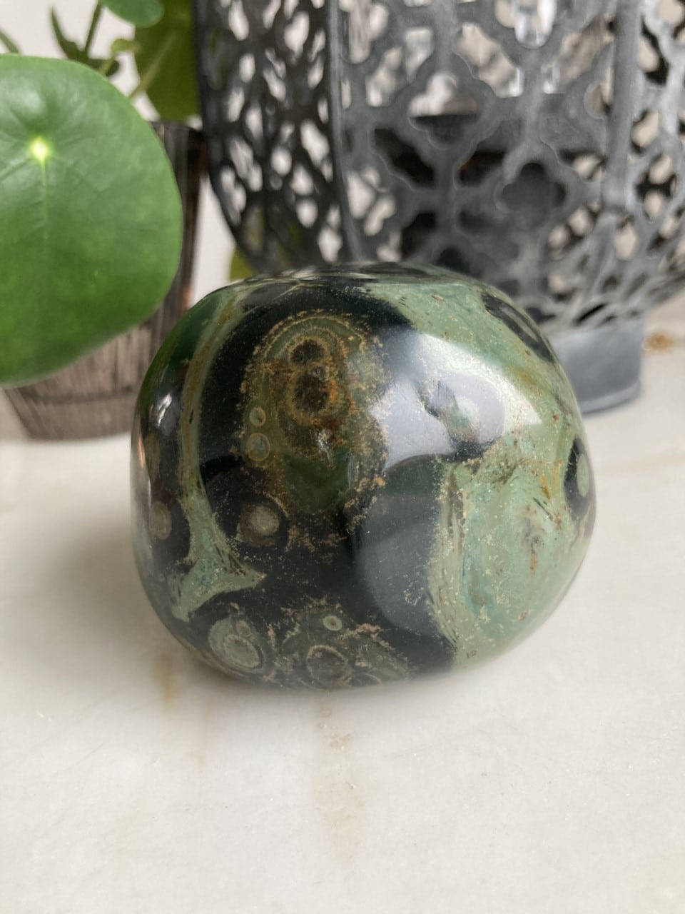 Rund, poleret Jaspis Kabamba Jumbo-sten med grønne og sorte mønstre foran en dekorativ lanternelignende genstand.