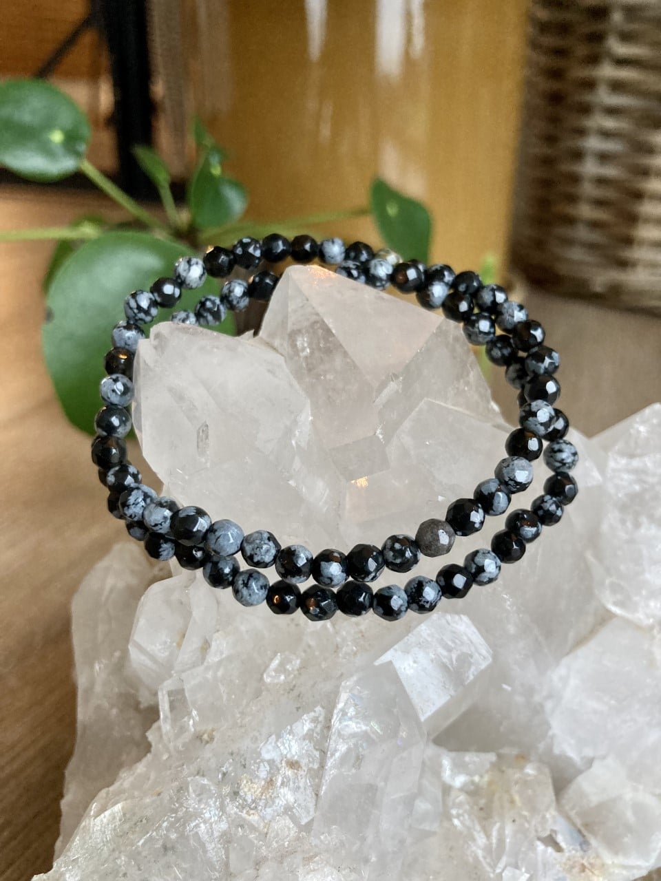 Obsidian Snefnug armbånd på en klynge af klare krystaller, med planter og kurv i baggrunden.