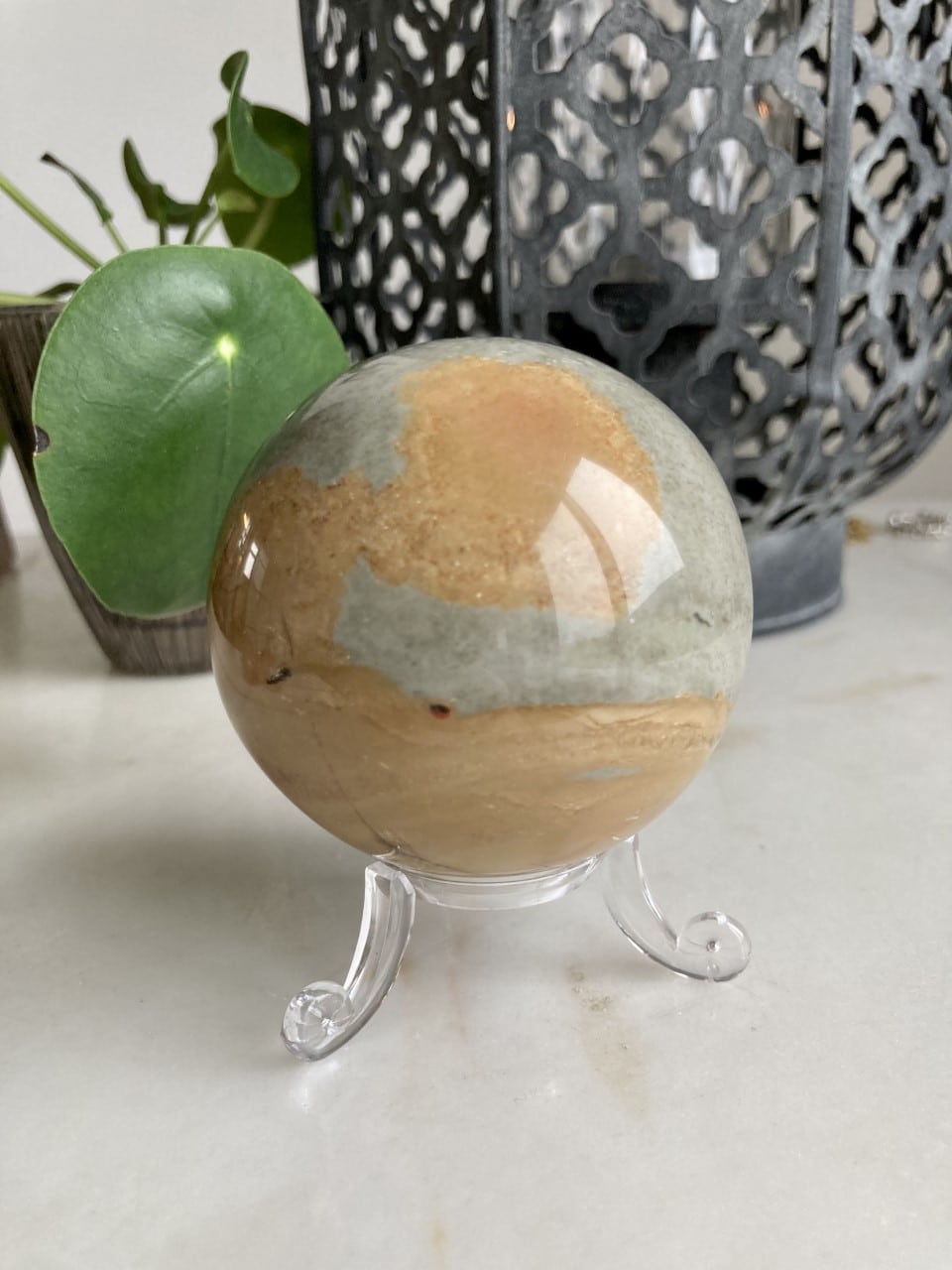 Jaspis Polychrome kugle med komplekse mønstre på klar stander, grøn plante og smedejernslampe i baggrunden.