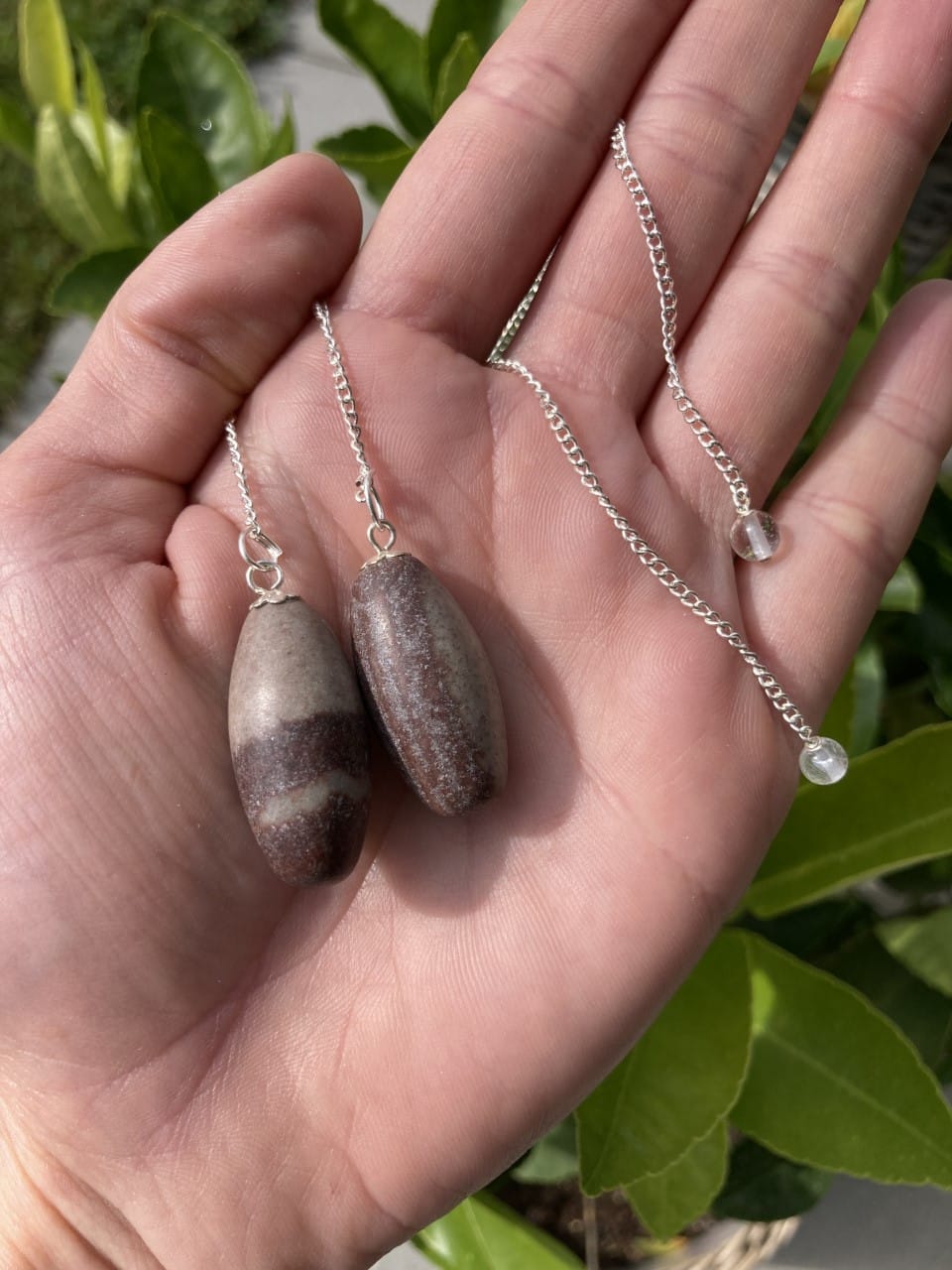 En hånd holder to Shiva Lingam penduler på sølvkæder mod en baggrund af grønne blade.