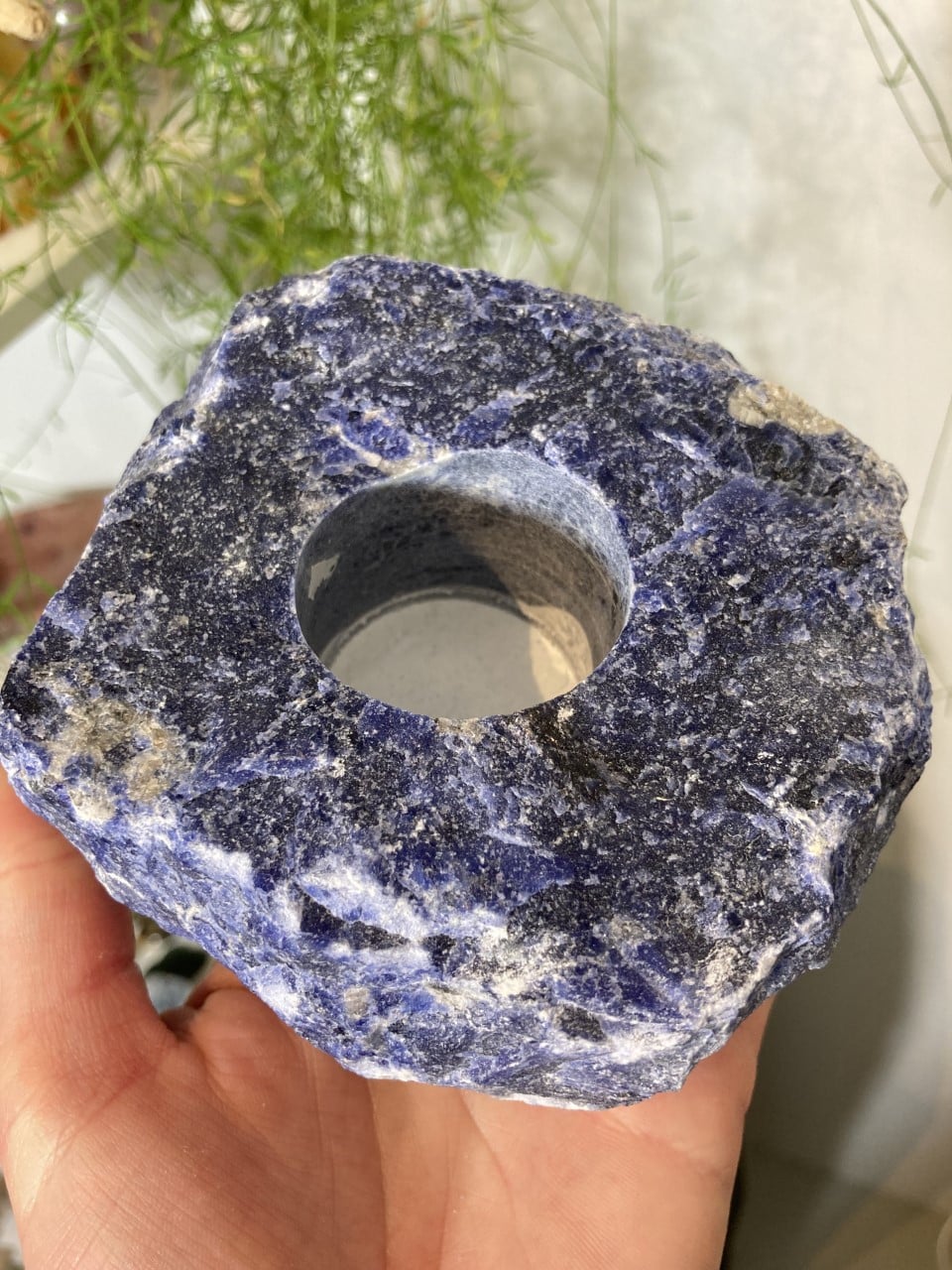 Sodalit fyrfadsstage nr. 3 i blå sten holdes i en hånd, omgivet af grønne planter i baggrunden.