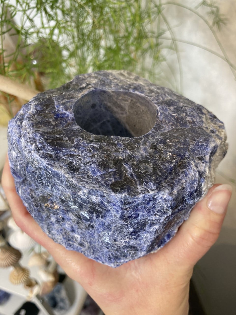 Sodalit fyrfadsstage nr. 1 holdt i en hånd, med blå og sorte nuancer, omgivet af grønne planter i baggrunden.