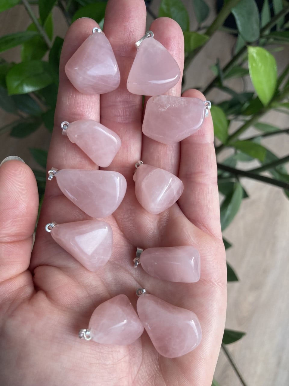 Håndfuld rosakvarts vedhæng i forskellige former, med grønne planter i baggrunden.