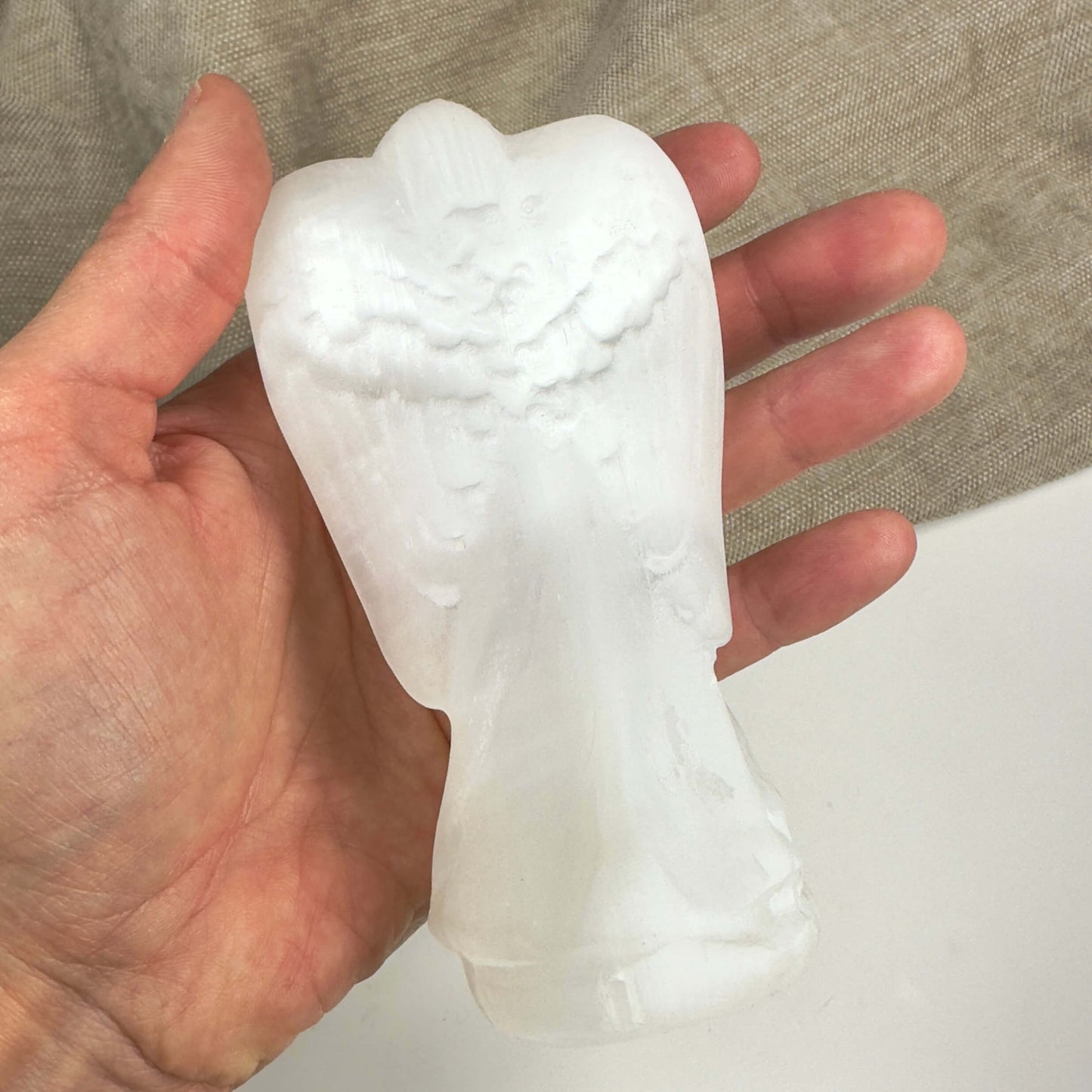 Hånd der holder en skulptur af en selenit engel på en beige baggrund.