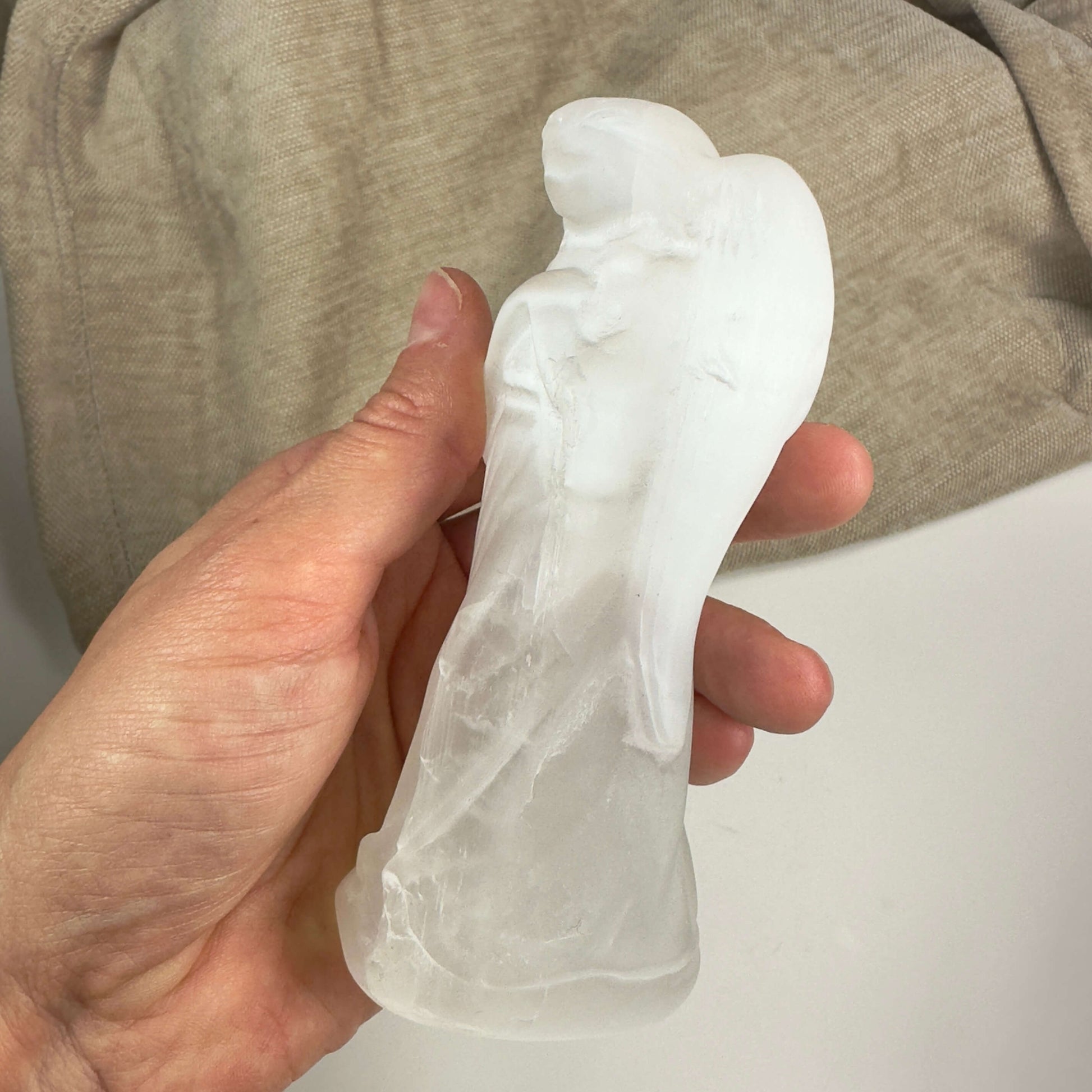 Hånd holder en selenit engel figur mod en beige baggrund.