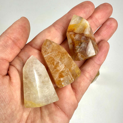 Hånd holder tre Golden healer tårne af kvarts, med gyldne og hvide nuancer, på en hvid baggrund.