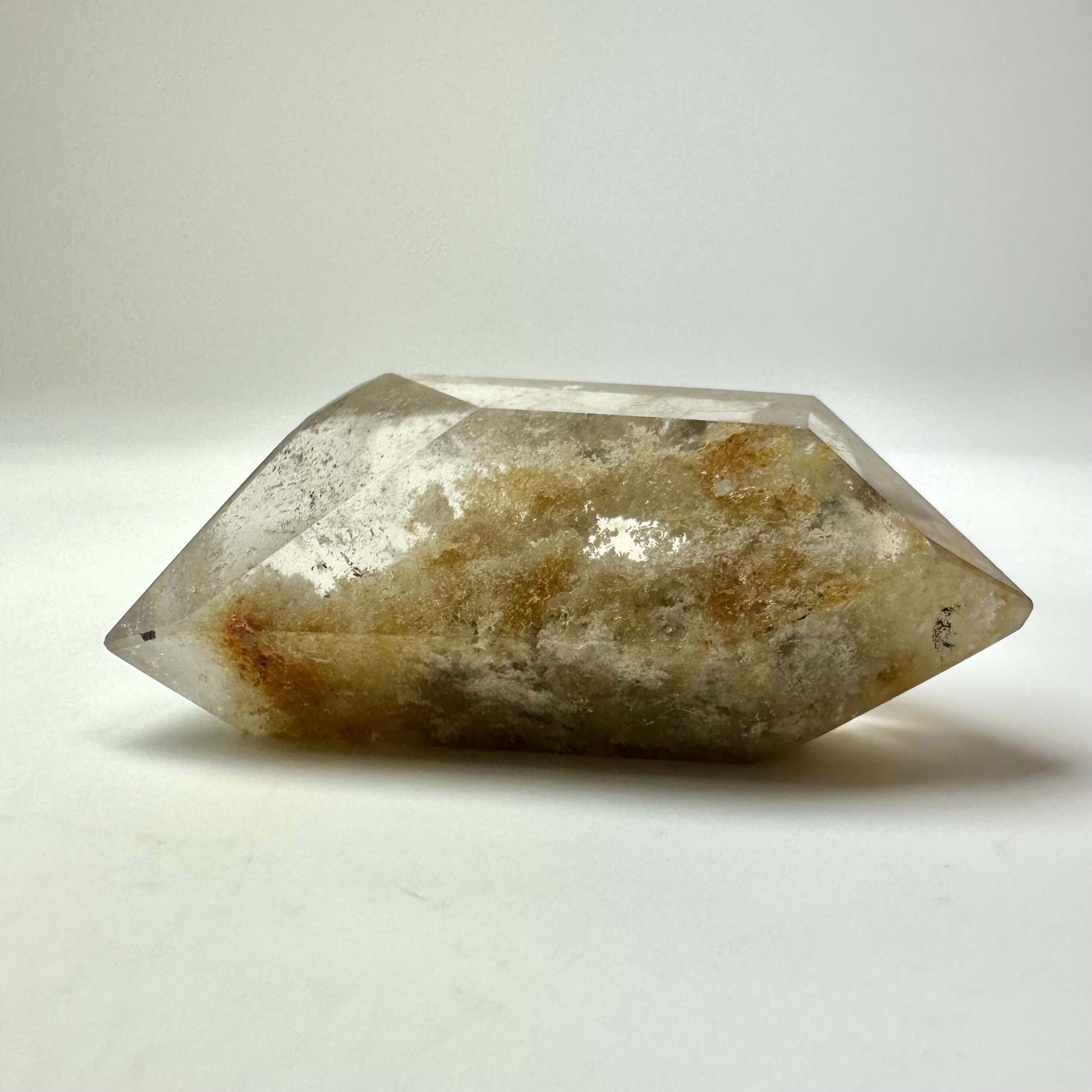 Fantomkvarts i fri form, placeret på en lys baggrund med synlige naturlige inkorporeringer og jordfarvede nuancer.