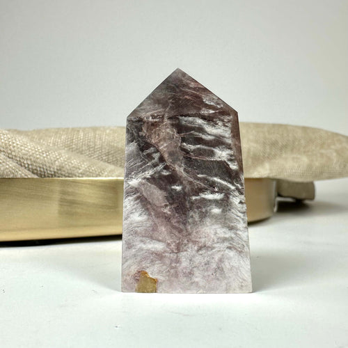 Lepidolit Mica-krystal i pyramideform, stående foran en beige baggrund på en hvid overflade.