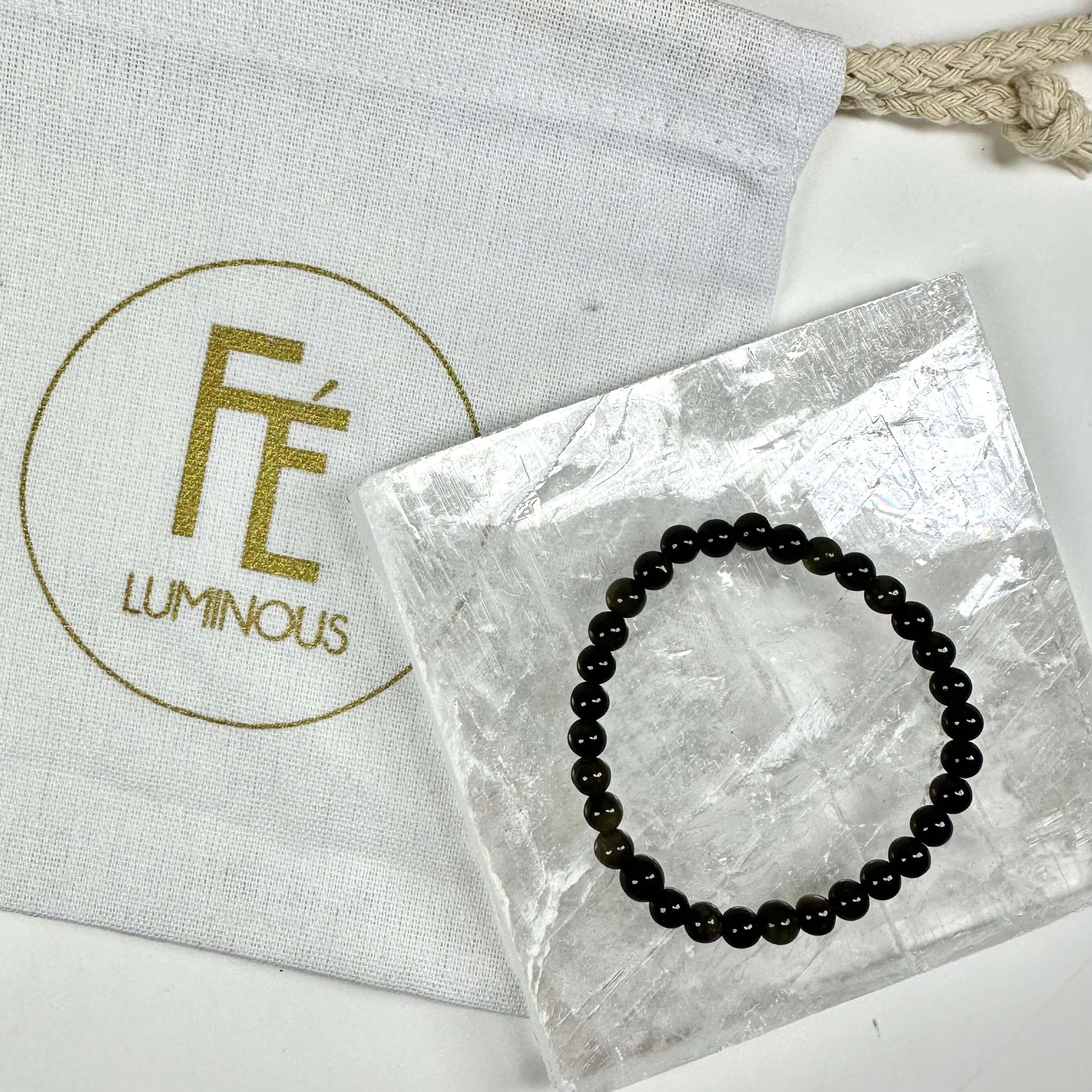 Armbånd opladningssæt inkl. gylden obsidian armbånd ved siden af en hvid pose med "FÉ LUMINOUS" logo.