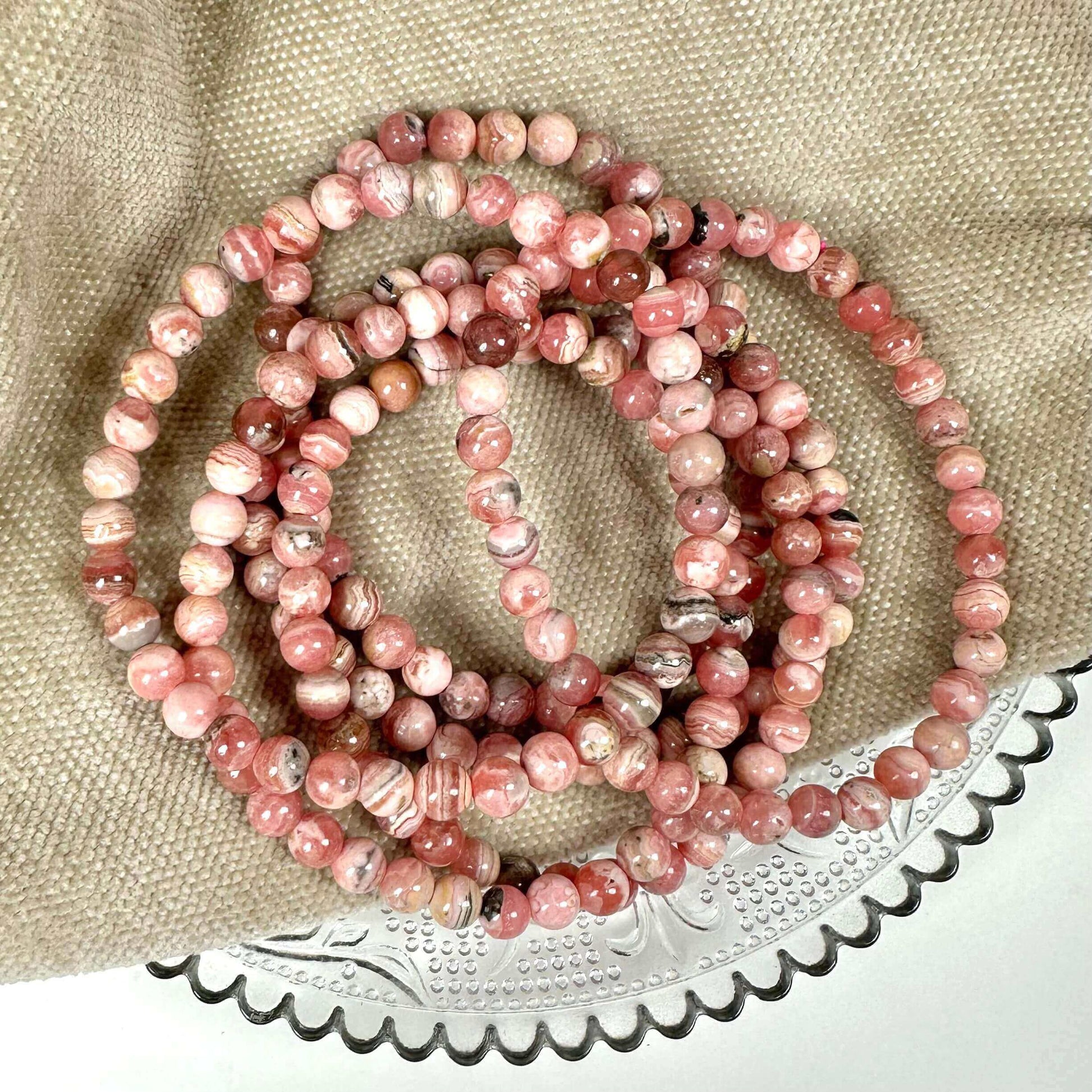 Lyserøde rhodochrosit perler på en glasplade med beige stofbaggrund.