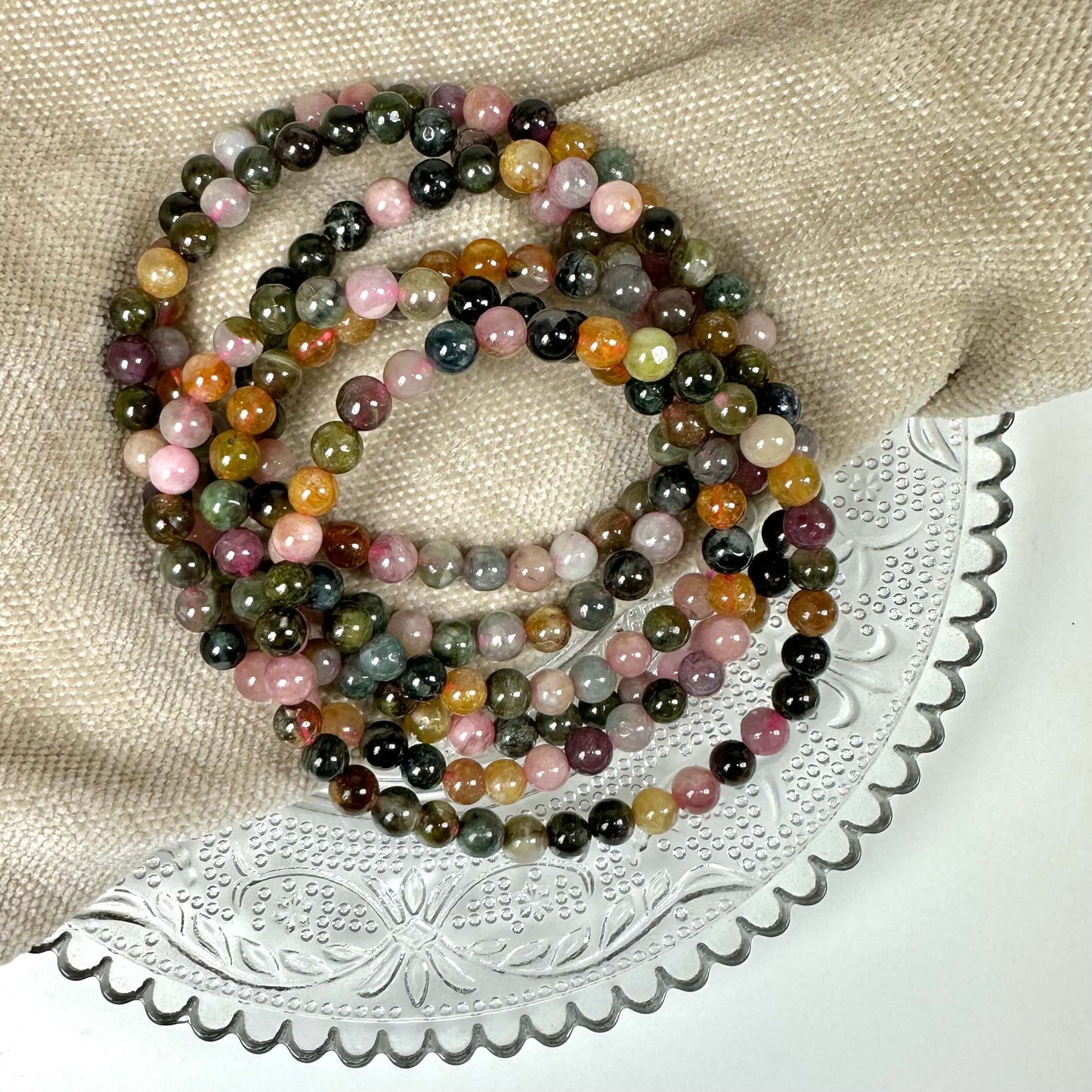Armbånd af farverige perler ligger på en beige klud og en dekoreret glasplade.