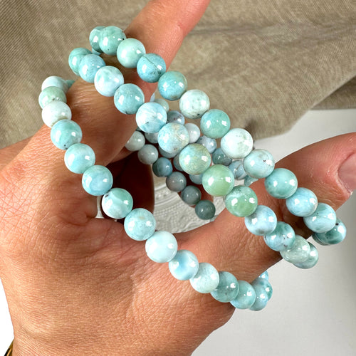 Hånd, der holder flere rækker af 6mm Larimar armbånd i lyseblå nuancer.