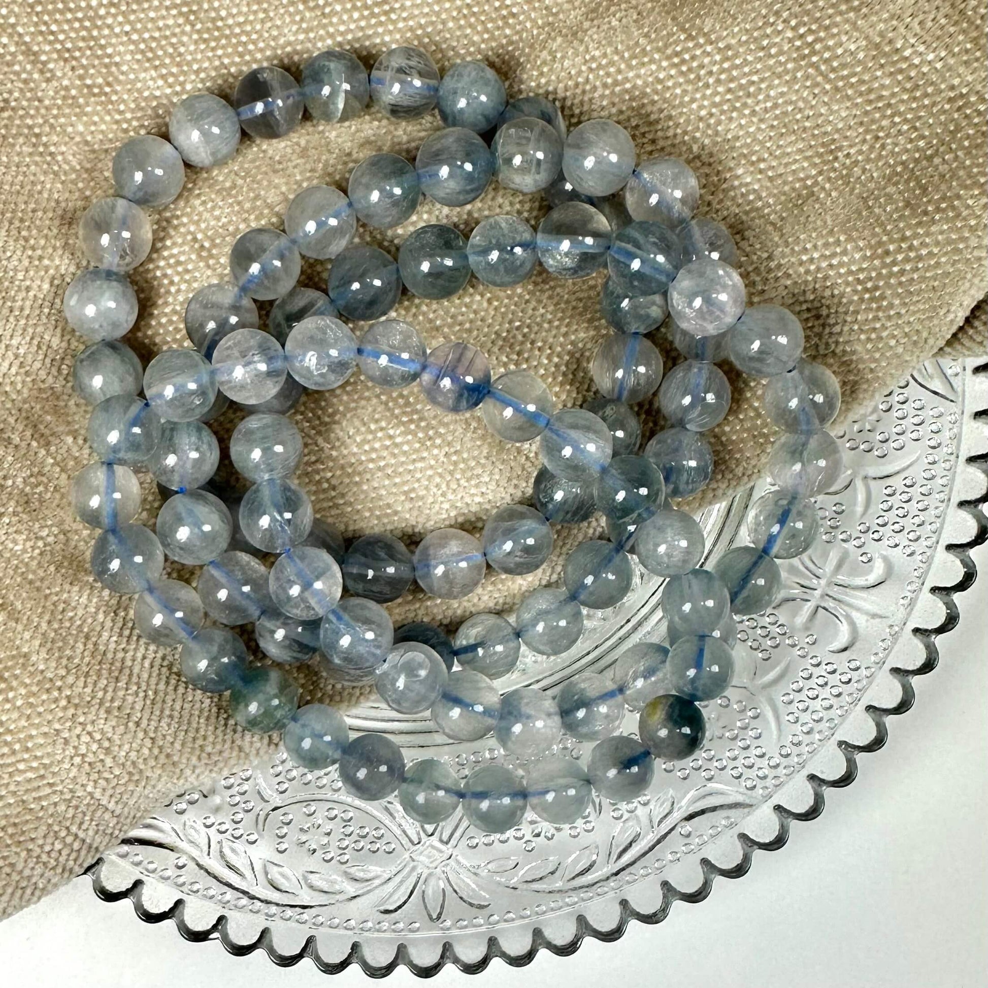 Blå fluorit armbånd 8mm ligger på en beige tekstil og dekoreret metalbakke.