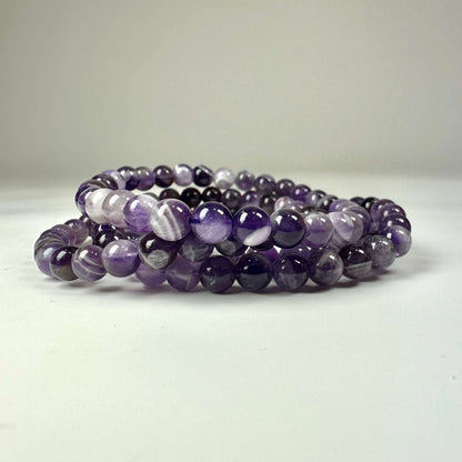 Ametyst Chevron armbånd 6mm med perler i nuancer af lilla og hvid, elegant placeret på en lys baggrund.